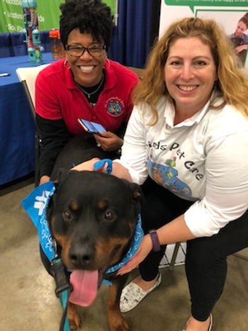 Two women smiling with a large Rottweiler wearing a blue bandana at an indoor event.