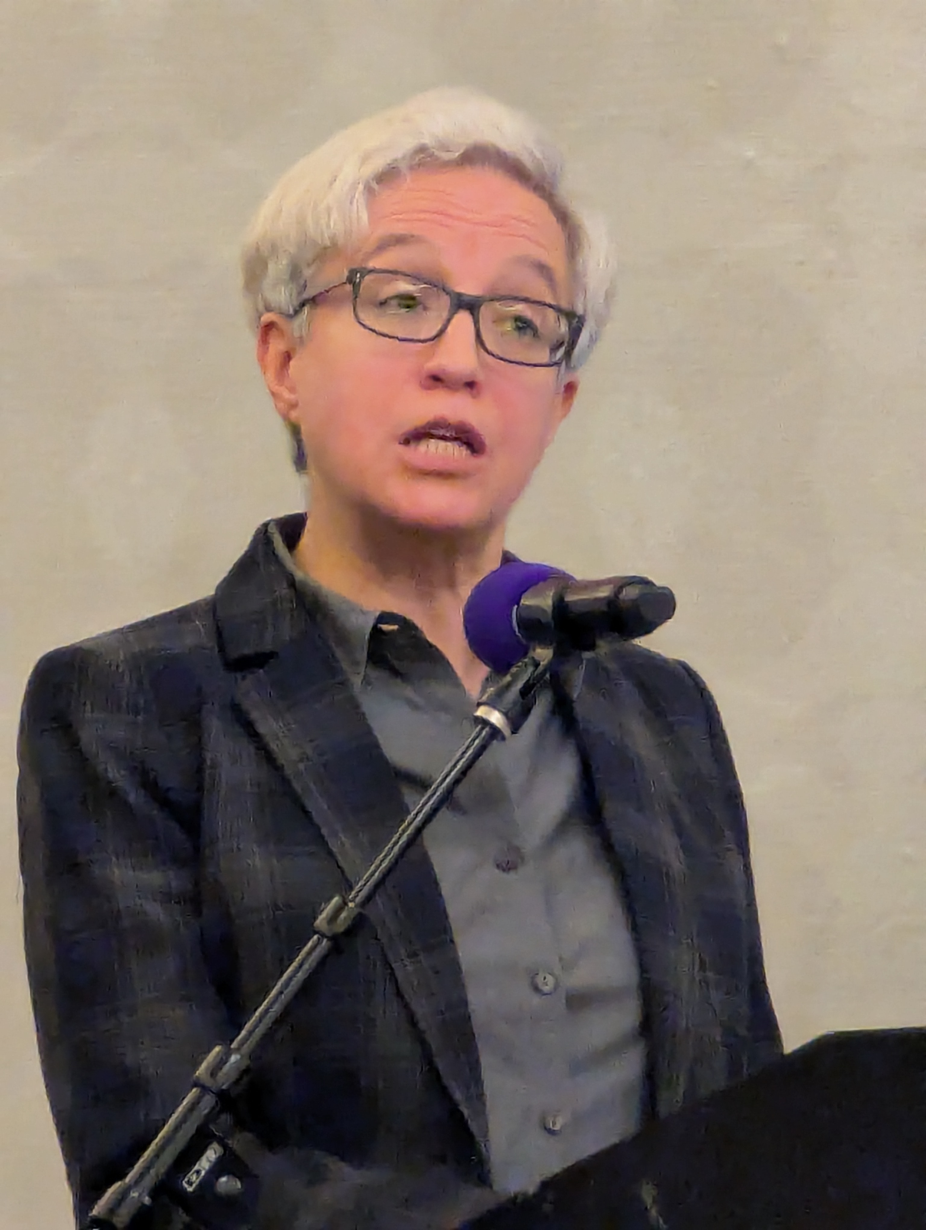 A person with short white hair and glasses speaks at a microphone podium.
