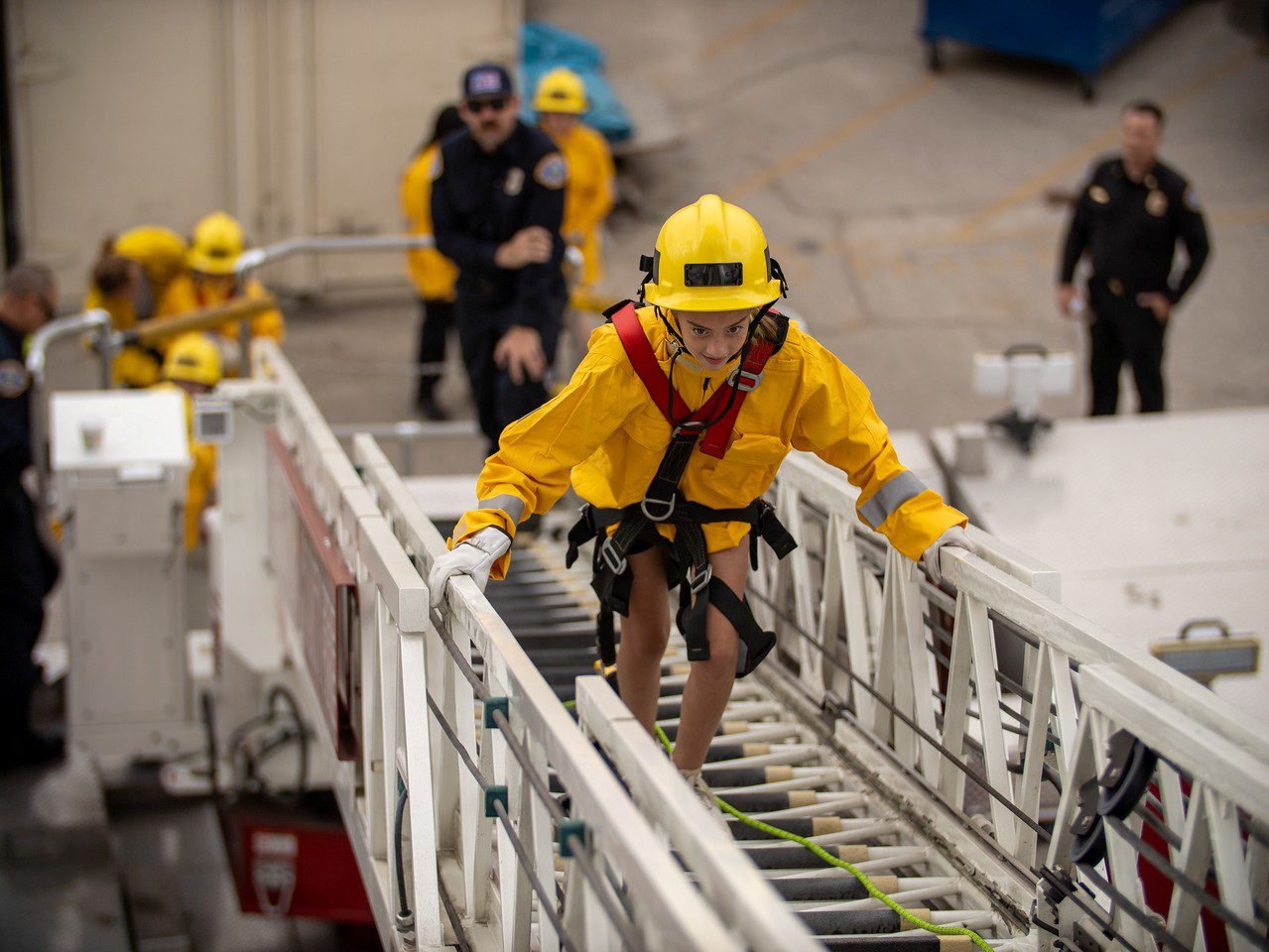 A young person in a yellow helmet and red harness climbs a fire truck aerial ladder.