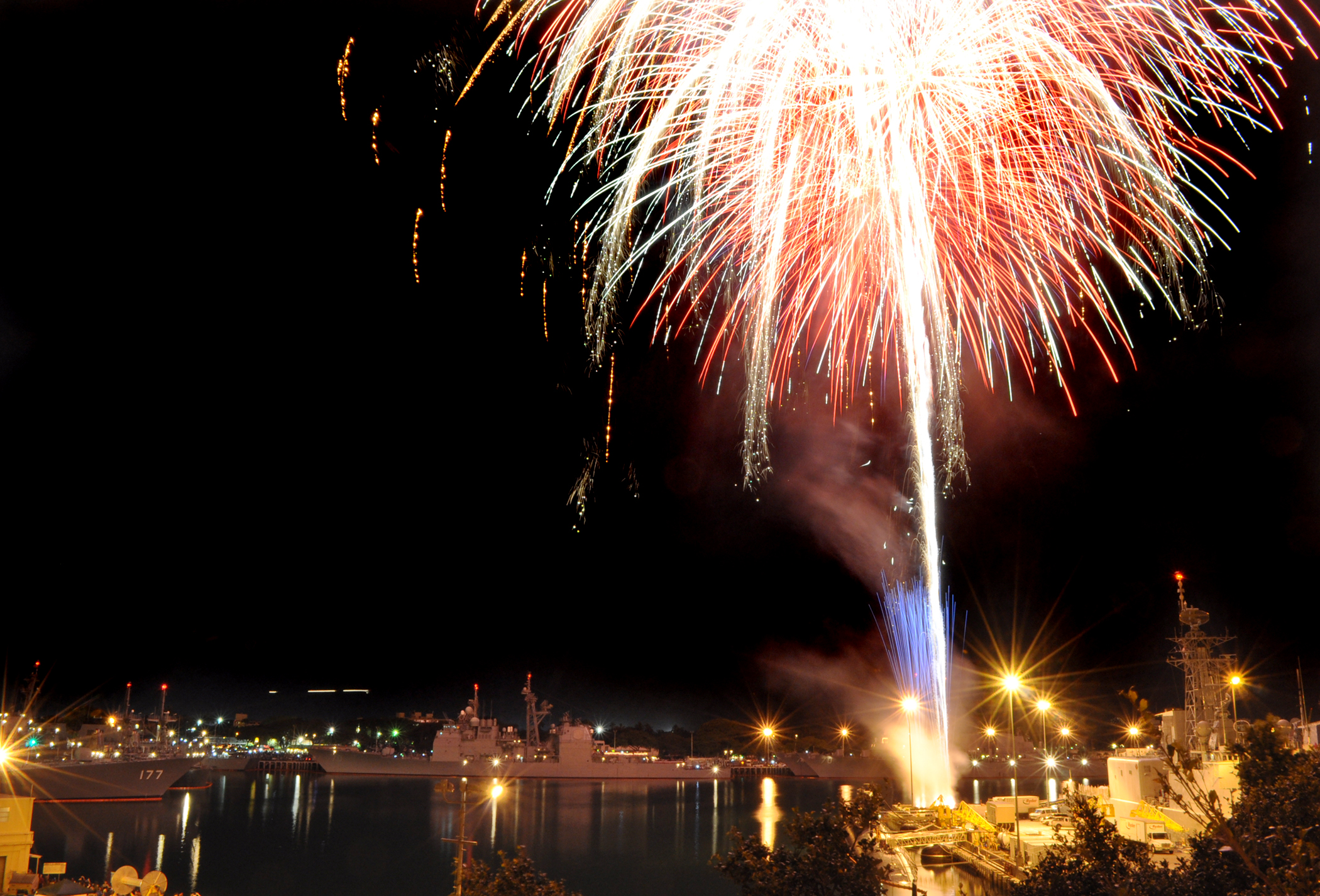 Fireworks bursting over naval ships docked at a harbor at night.
