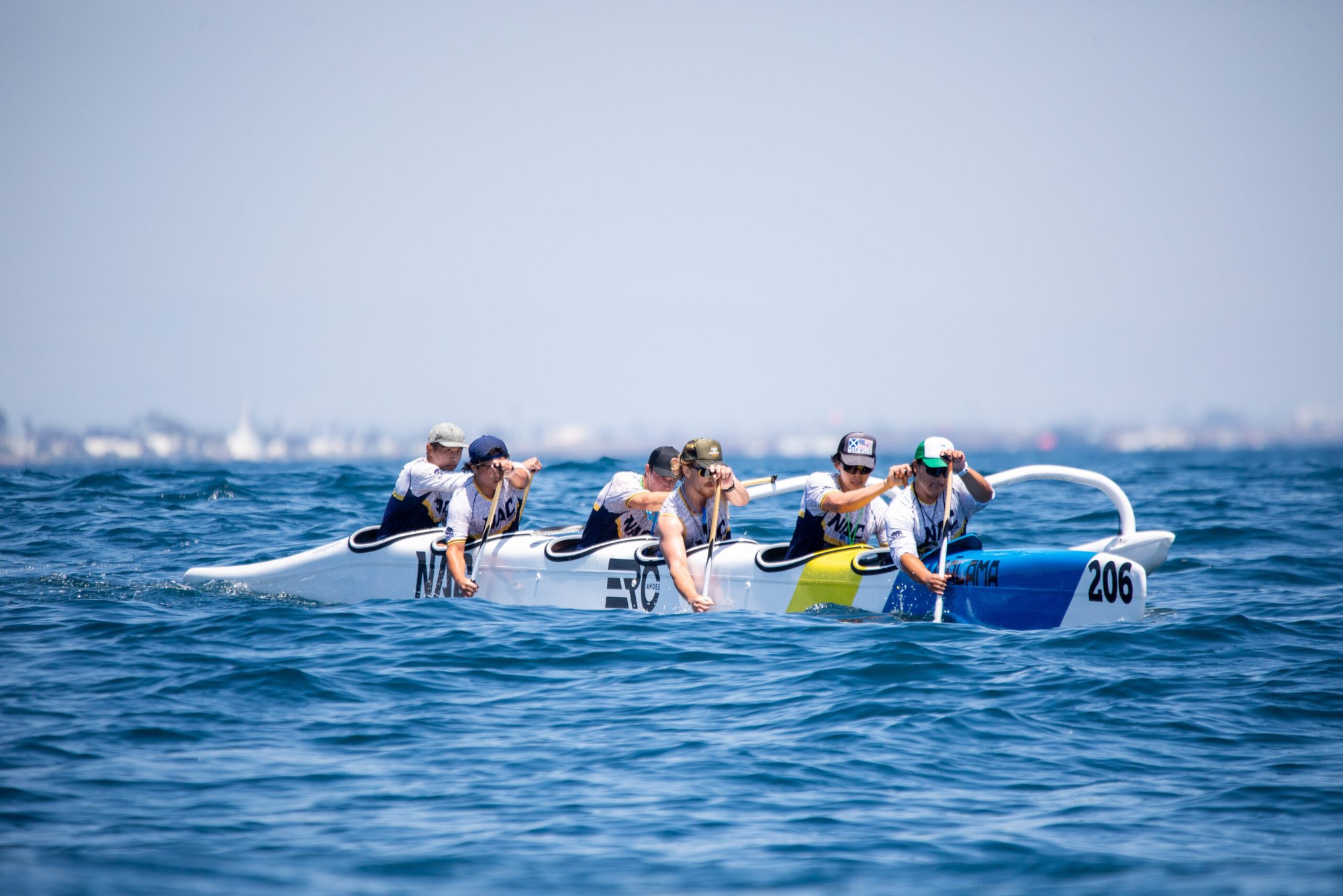 Six paddlers racing a yellow and white outrigger canoe numbered 206 on open ocean.