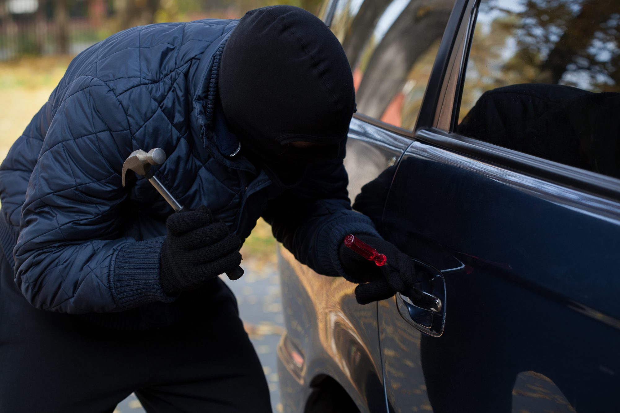 A masked burglar trying to break in a car using a hammer