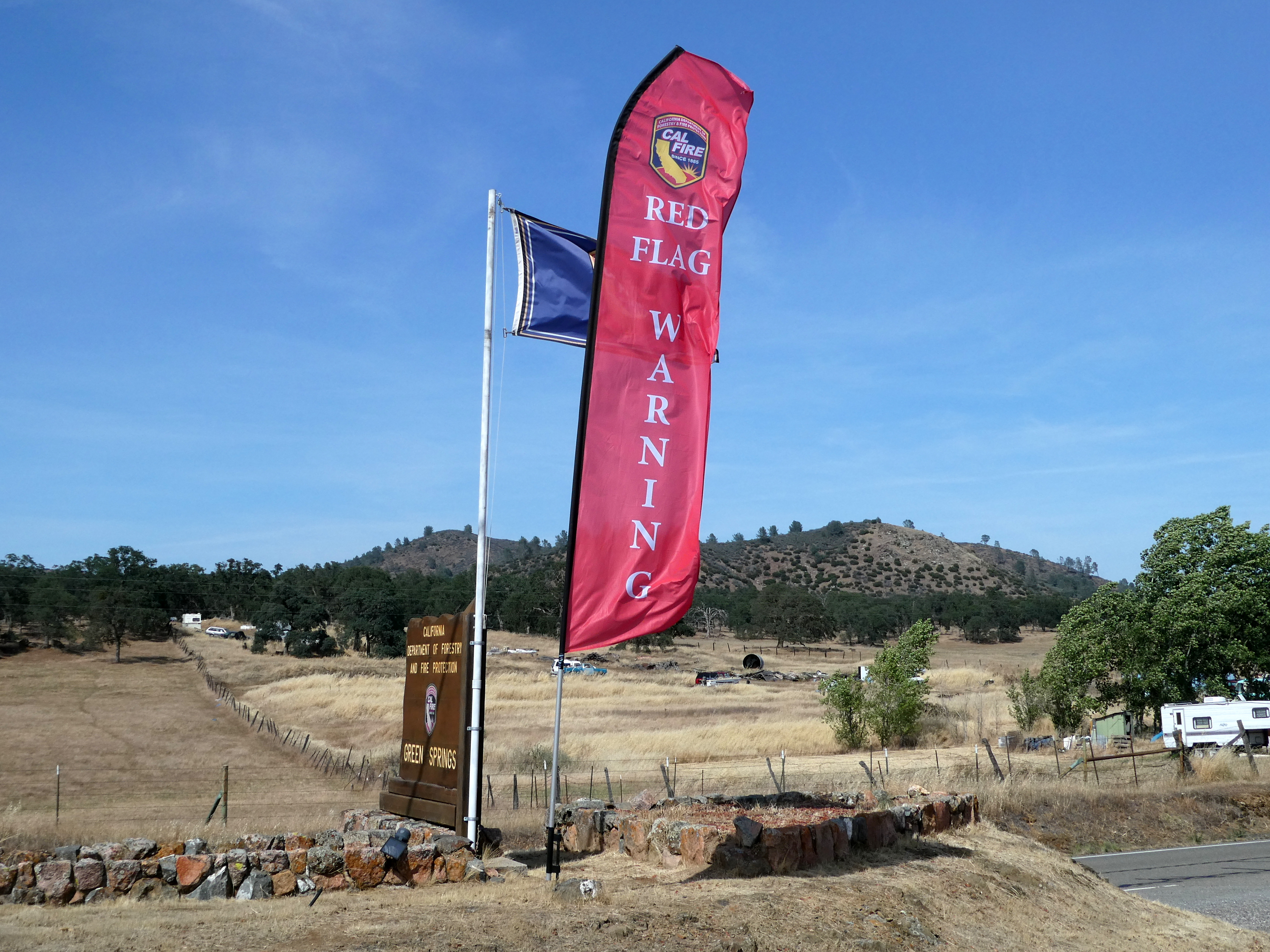 Tall red feather banner reading Red Flag Warning on a pole, dry hills behind.