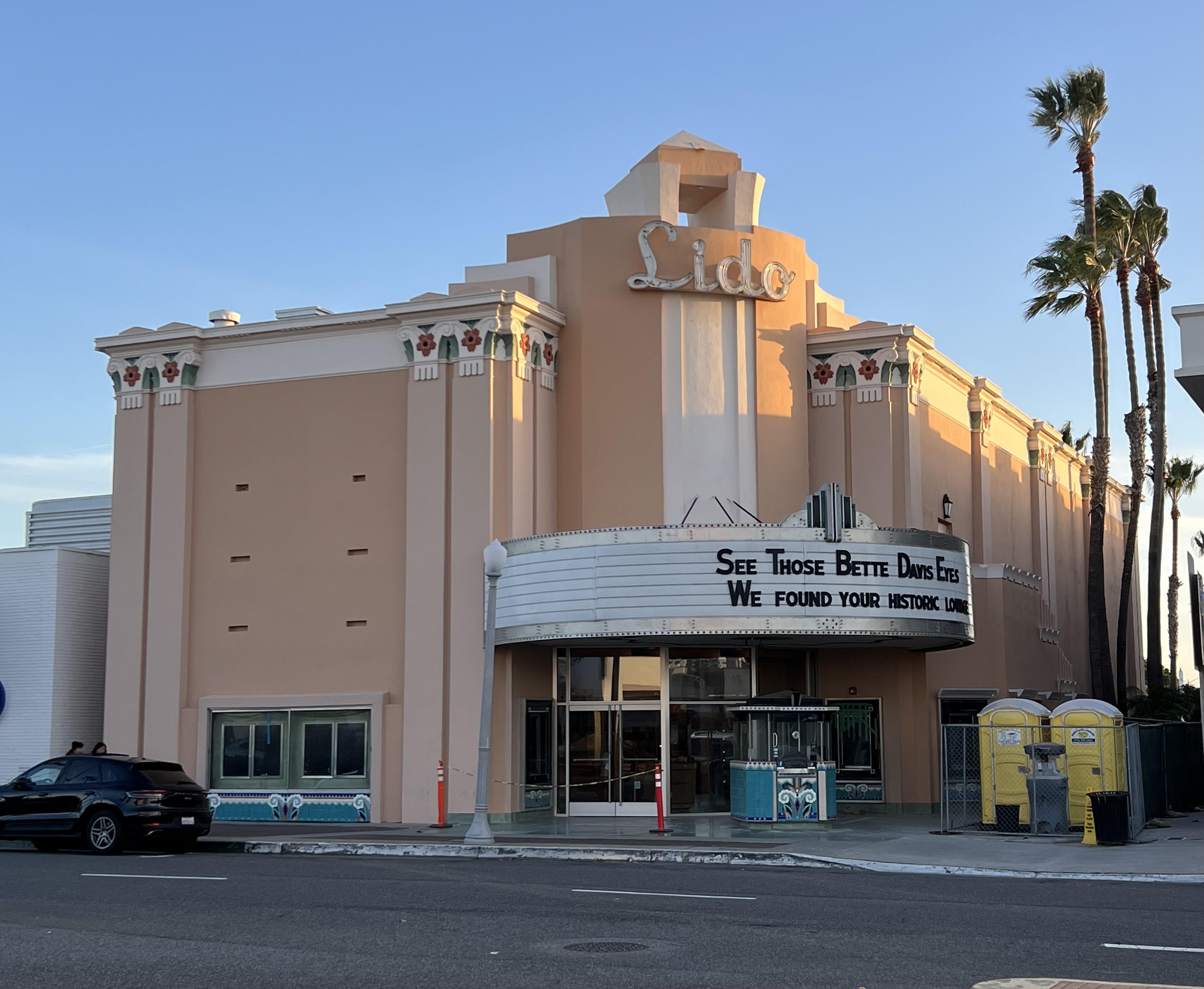 Art Deco facade of the Lido Theater with neon sign, marquee, and palm trees on a sunny day.