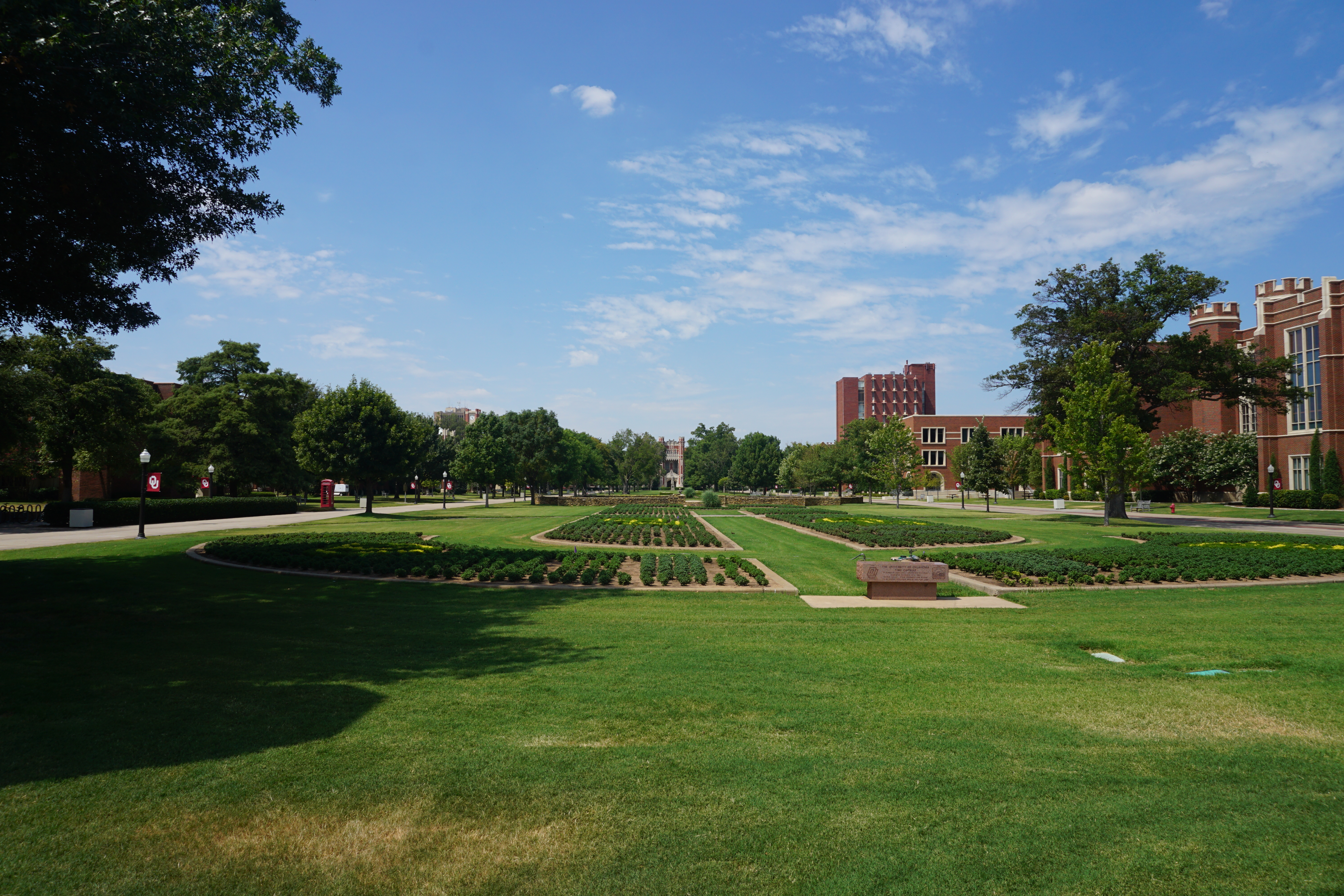 Wide green lawn with garden beds and walkway flanked by trees and red-brick university buildings.