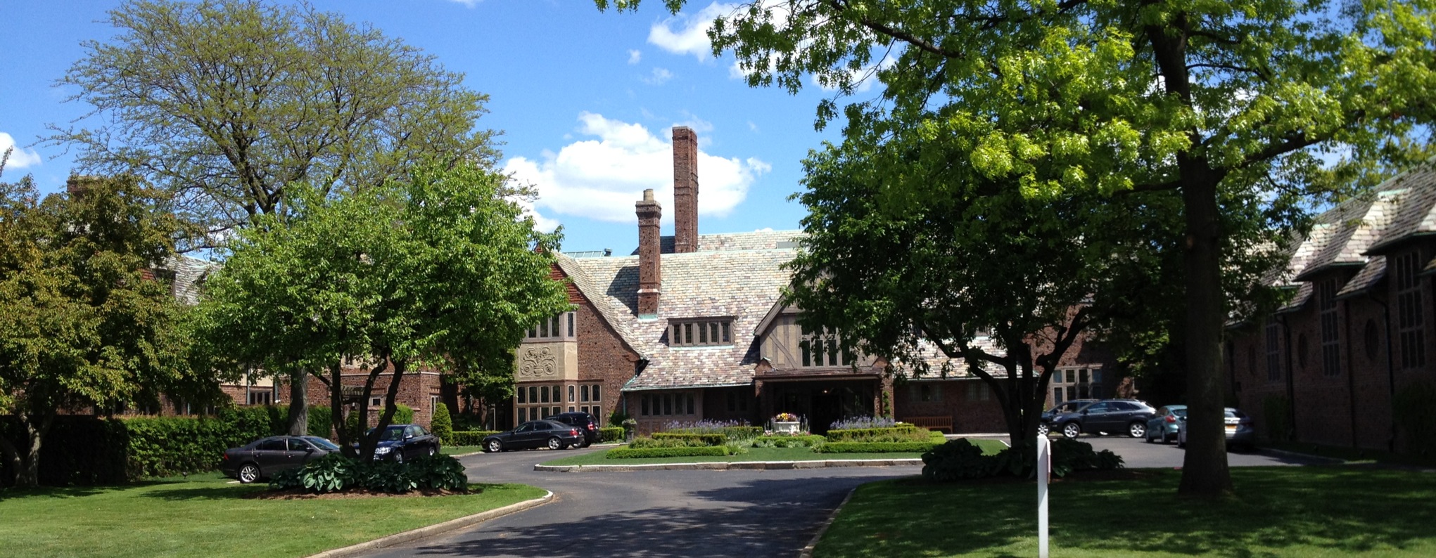 Large Tudor-style brick building with chimneys and a curved driveway lined with parked cars.