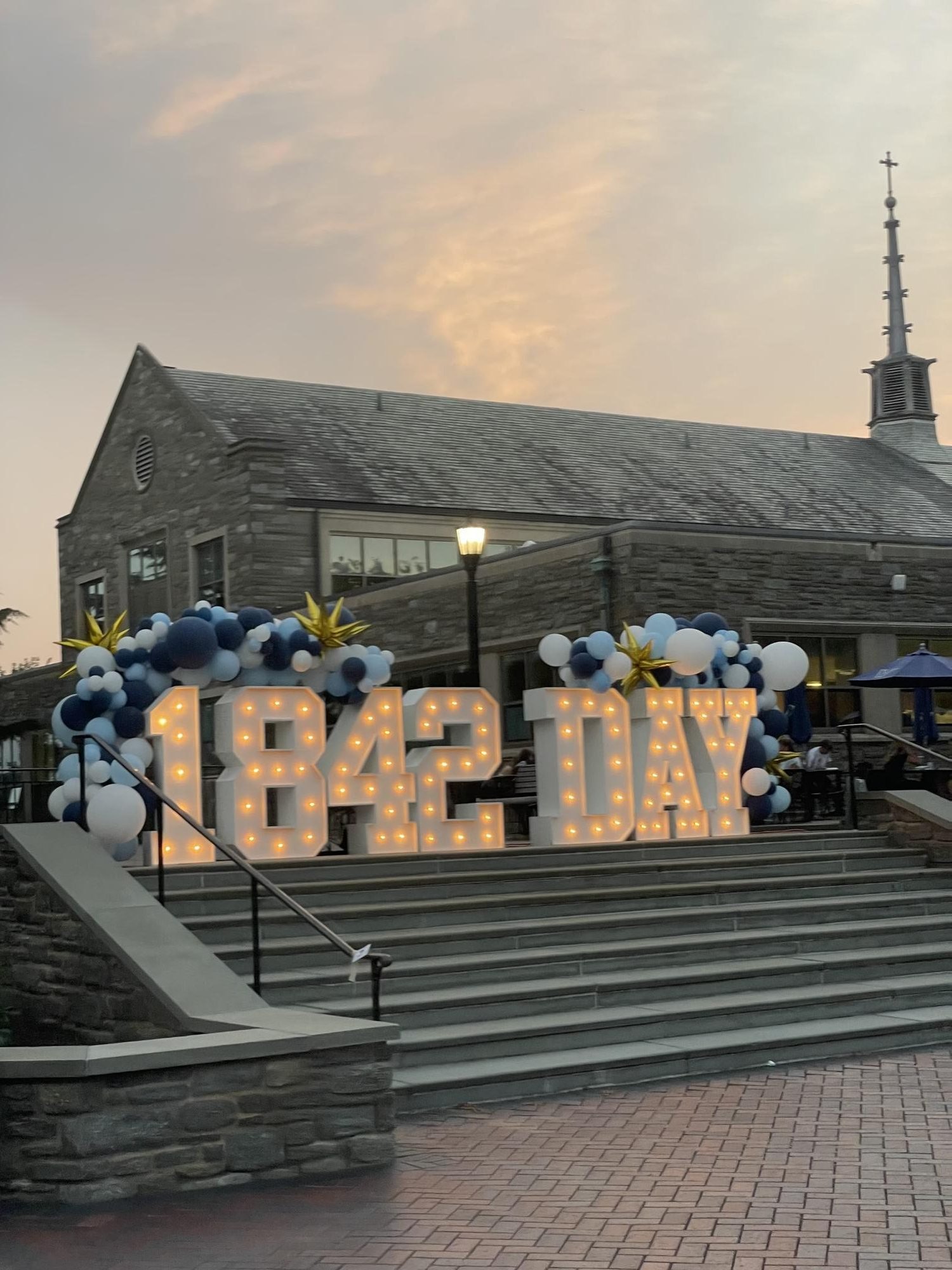 Illuminated marquee letters spelling '1842 DAY' surrounded by navy and gold balloons at dusk.