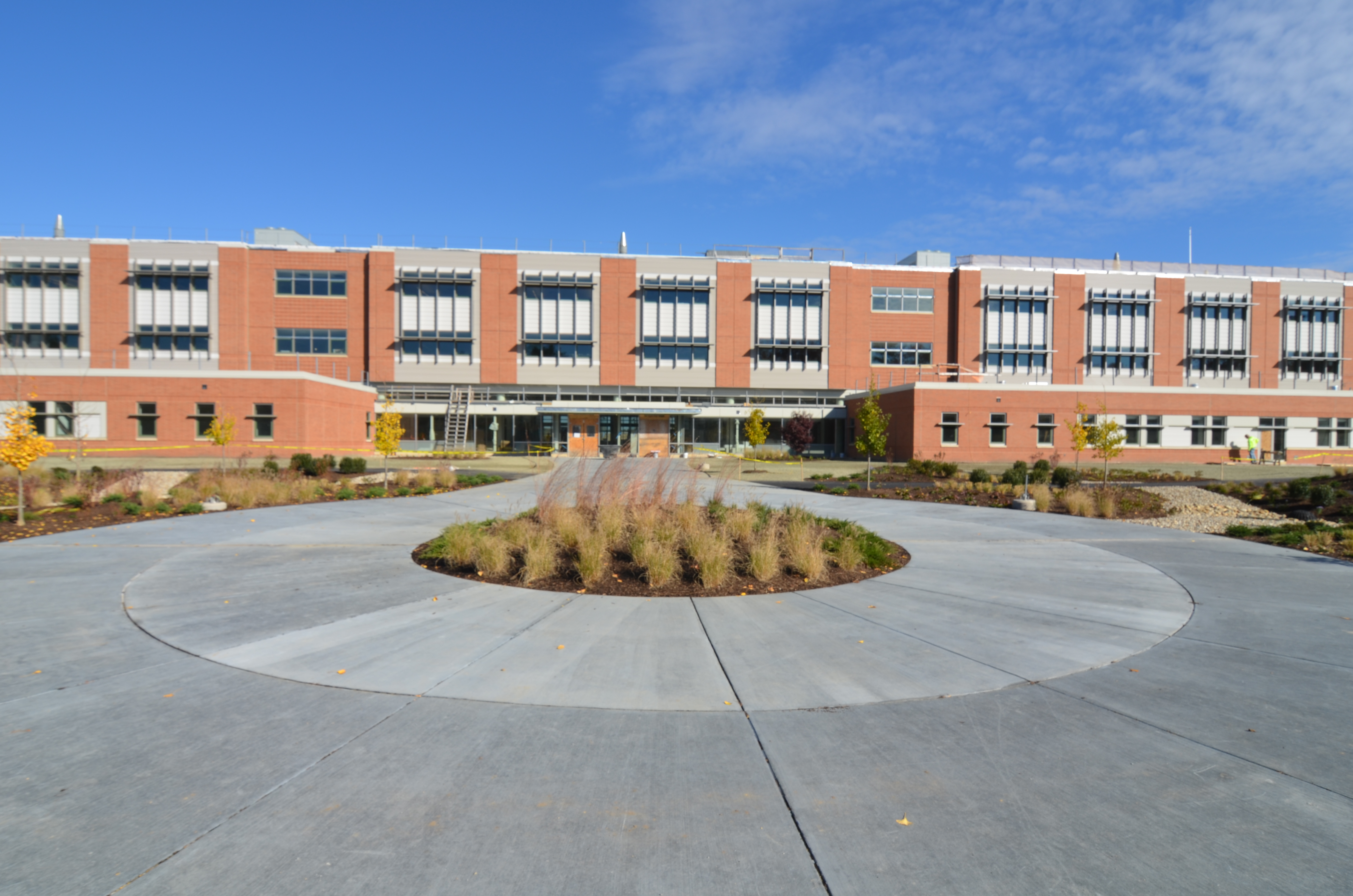 Modern multi-story brick school building with a circular concrete plaza and young trees in the foreground.