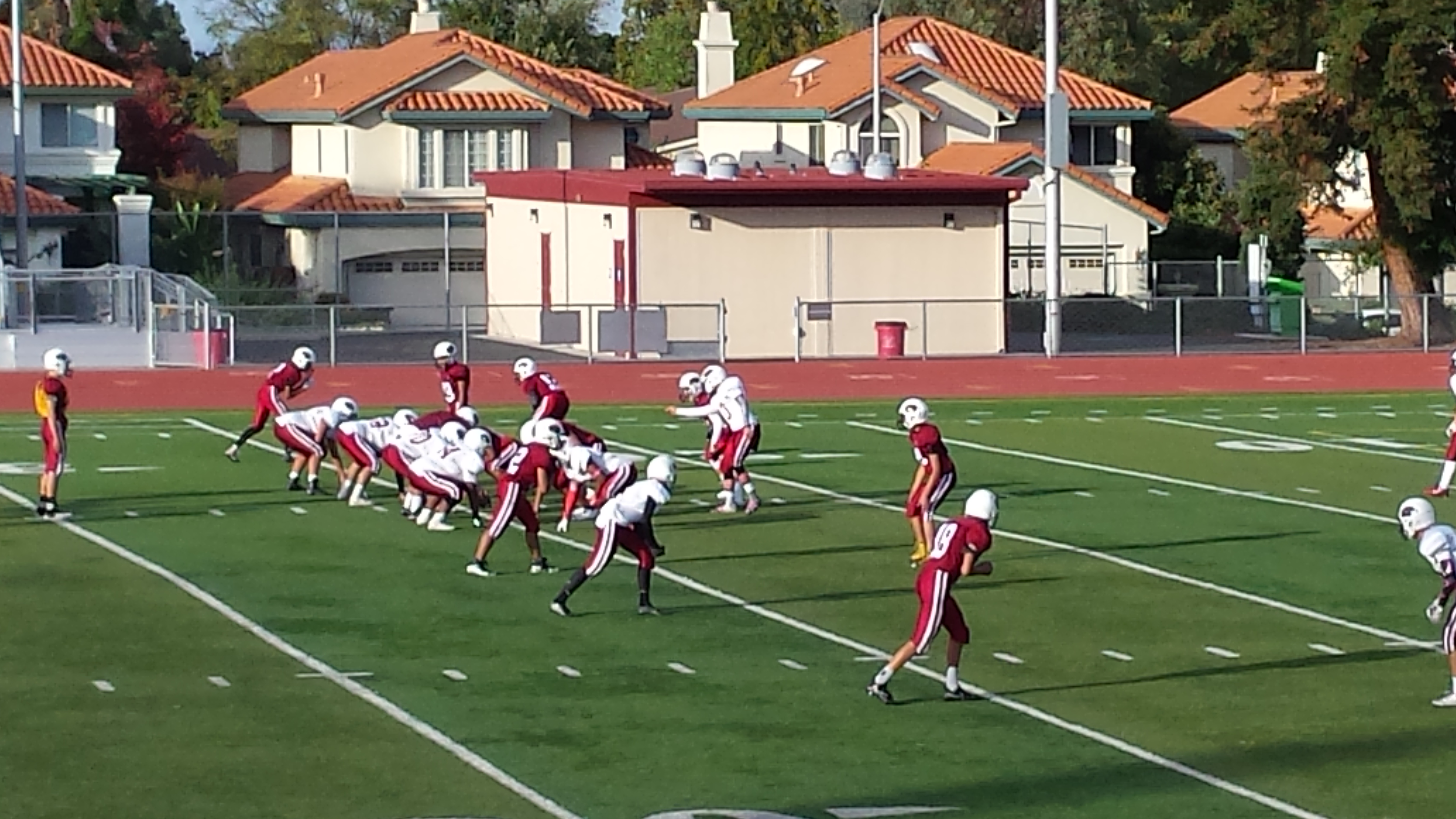 High school football players in red and white uniforms lined up at the line of scrimmage on a green turf field.