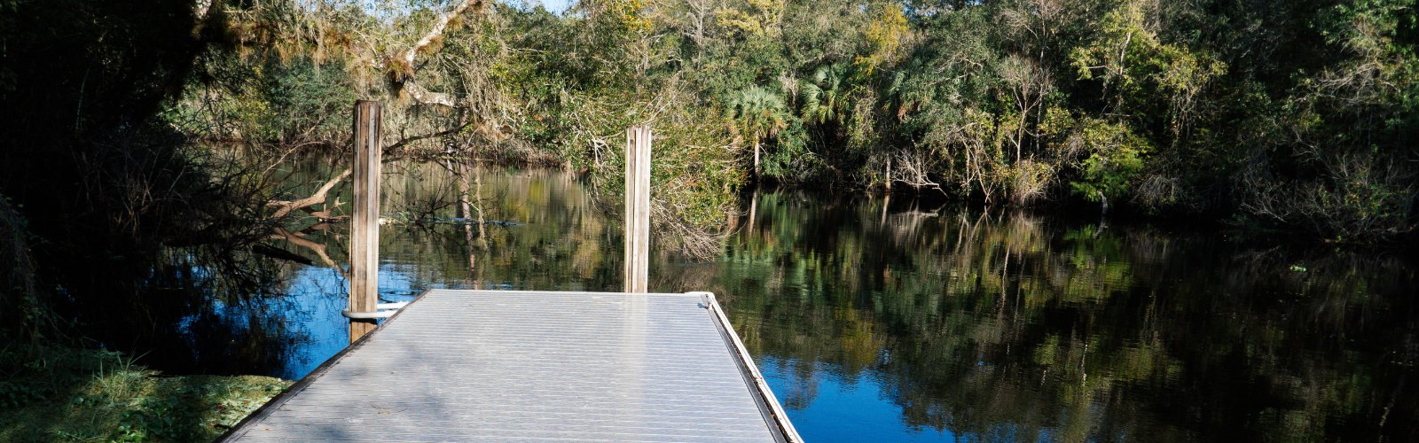 Wooden boardwalk with metal rails stretching over a calm, tree-lined river.