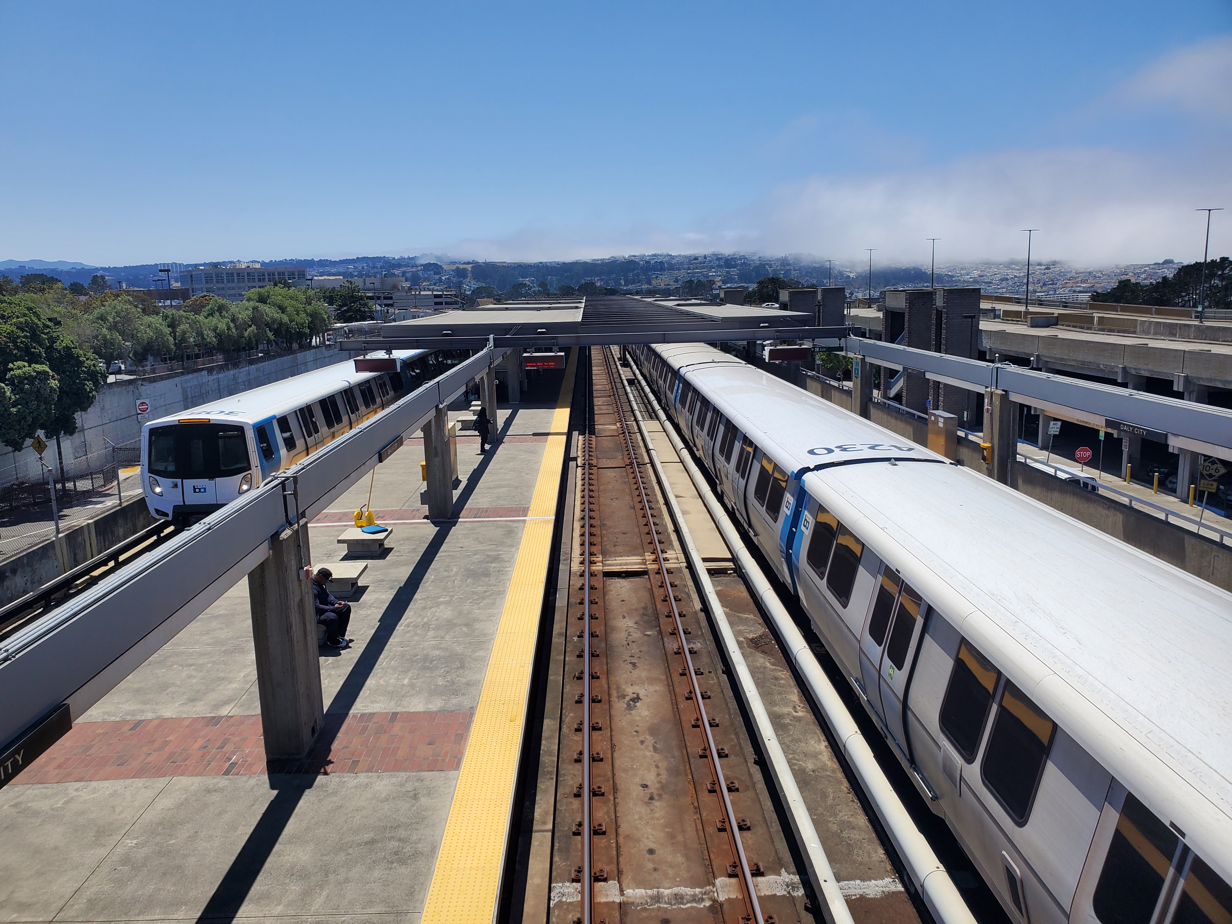 Two BART trains on parallel platforms at an elevated station with converging tracks.