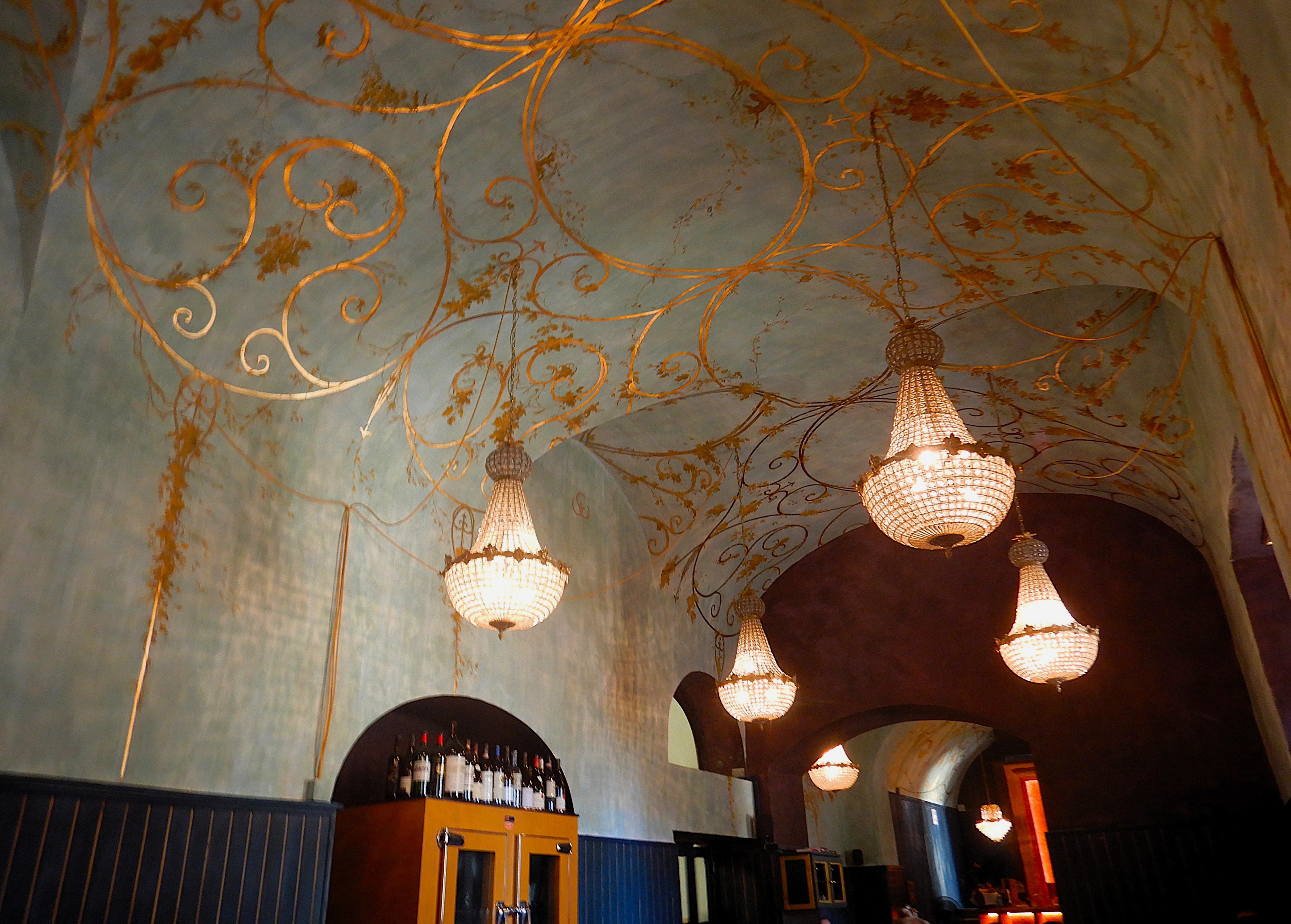 Ornate restaurant dining room with painted ceiling, crystal chandeliers, and a small bar.