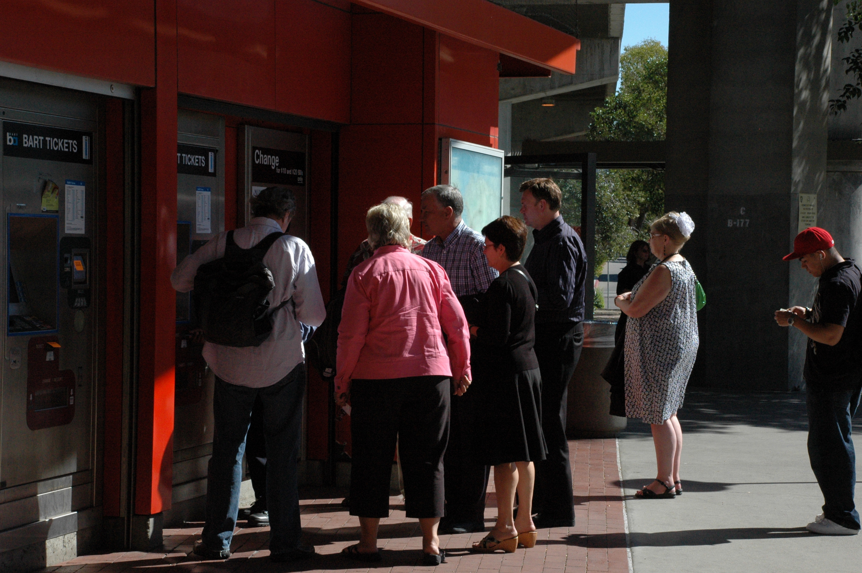 Several people standing in line at outdoor BART ticket vending machines on a red station wall.