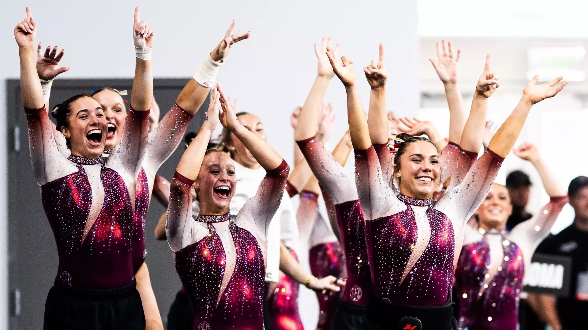 Group of female gymnasts in maroon rhinestone leotards cheering with arms raised.