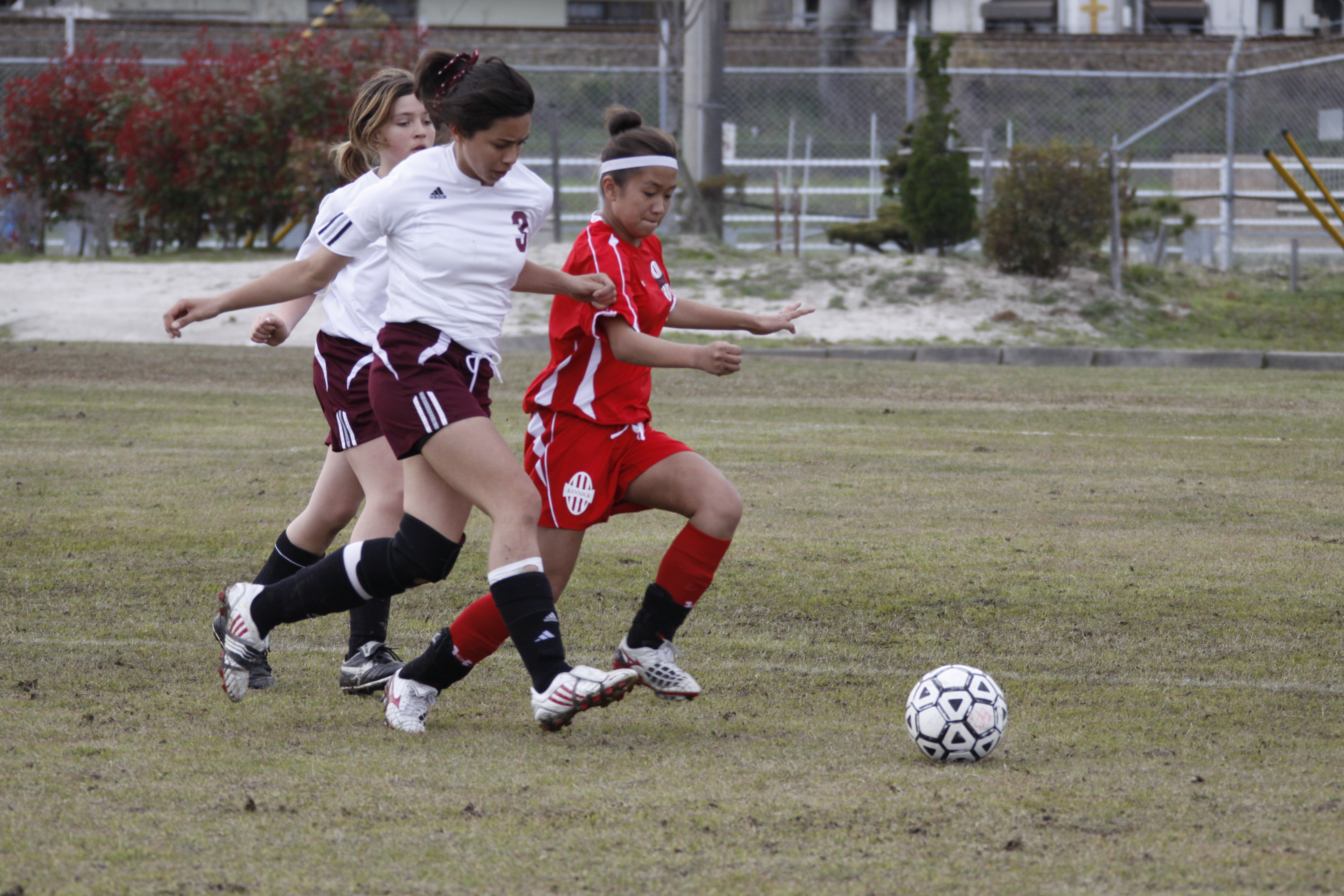 Three female soccer players racing to control a ball on a grass field.