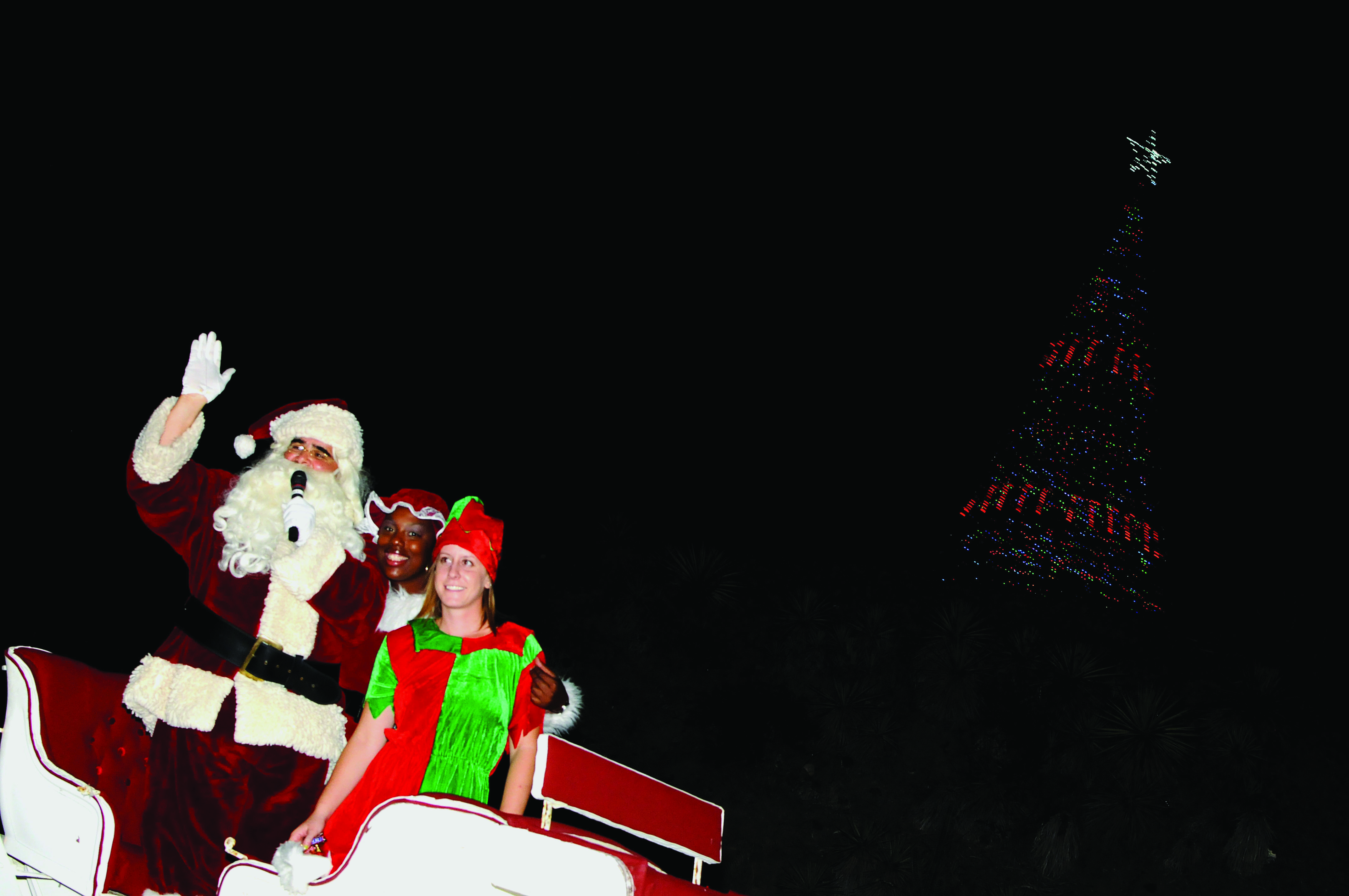 Santa Claus in red suit waves from a decorated parade float at night.