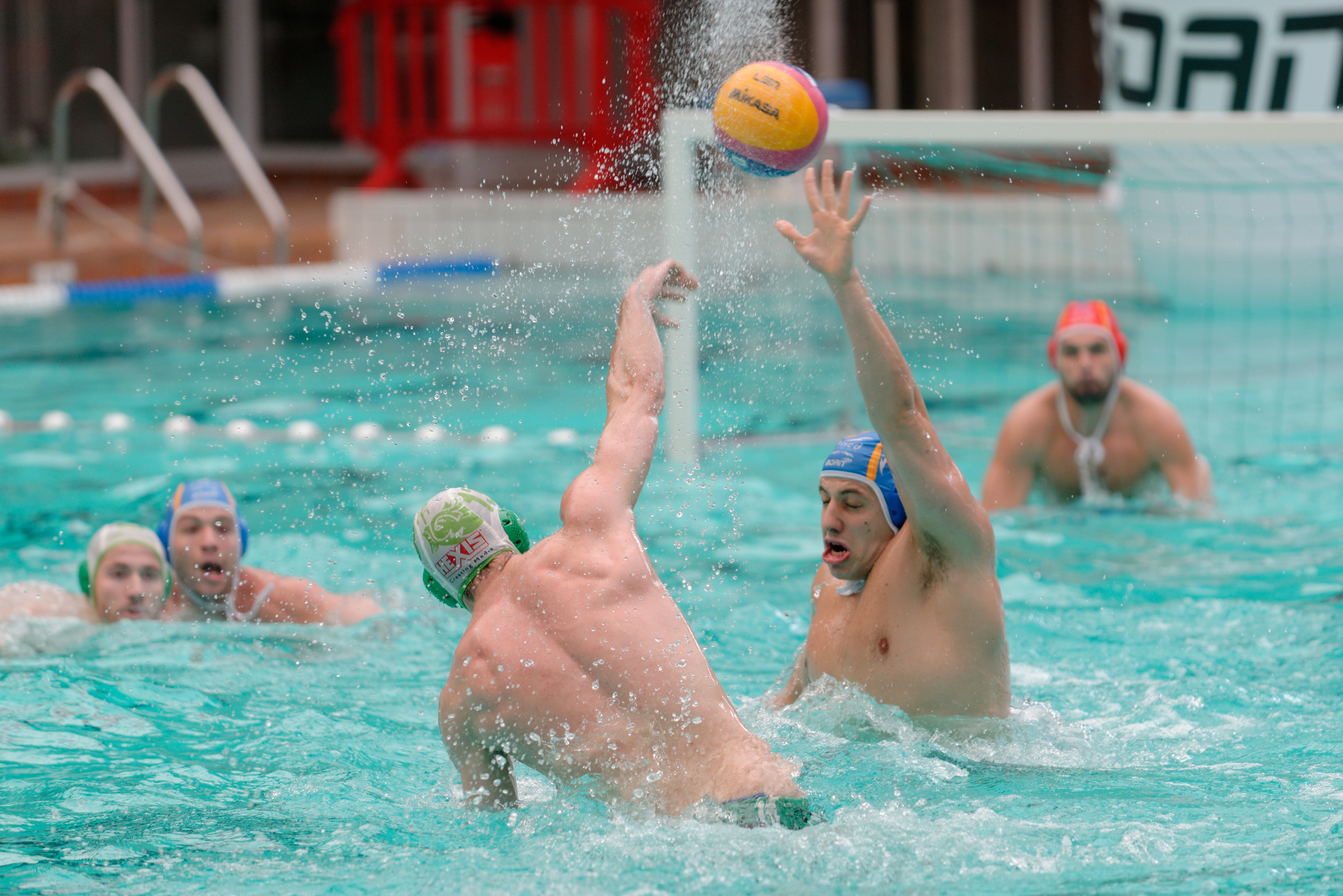 Male water polo players in a pool, one throwing the ball while another defends.