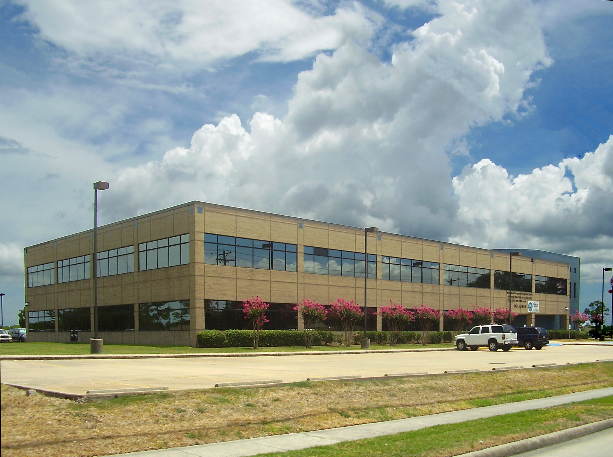 Two-story tan brick office building with large windows and pink flowering shrubs in front.