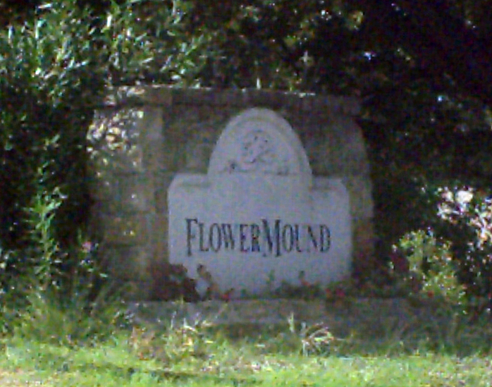 Stone monument sign reading 'Flower Mound' flanked by trees and shrubs.