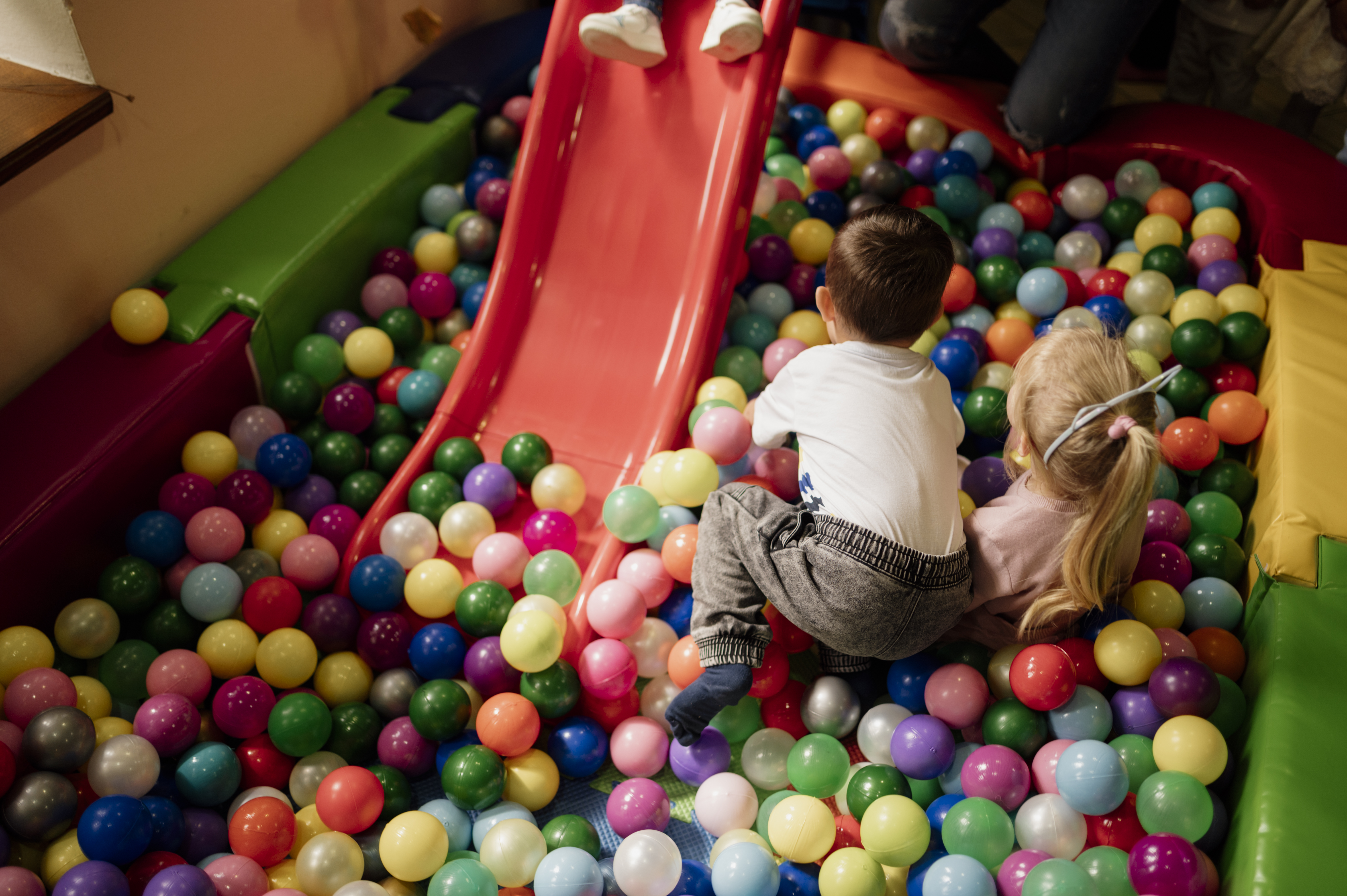 Two young children play in a colorful ball pit with a red slide behind them.