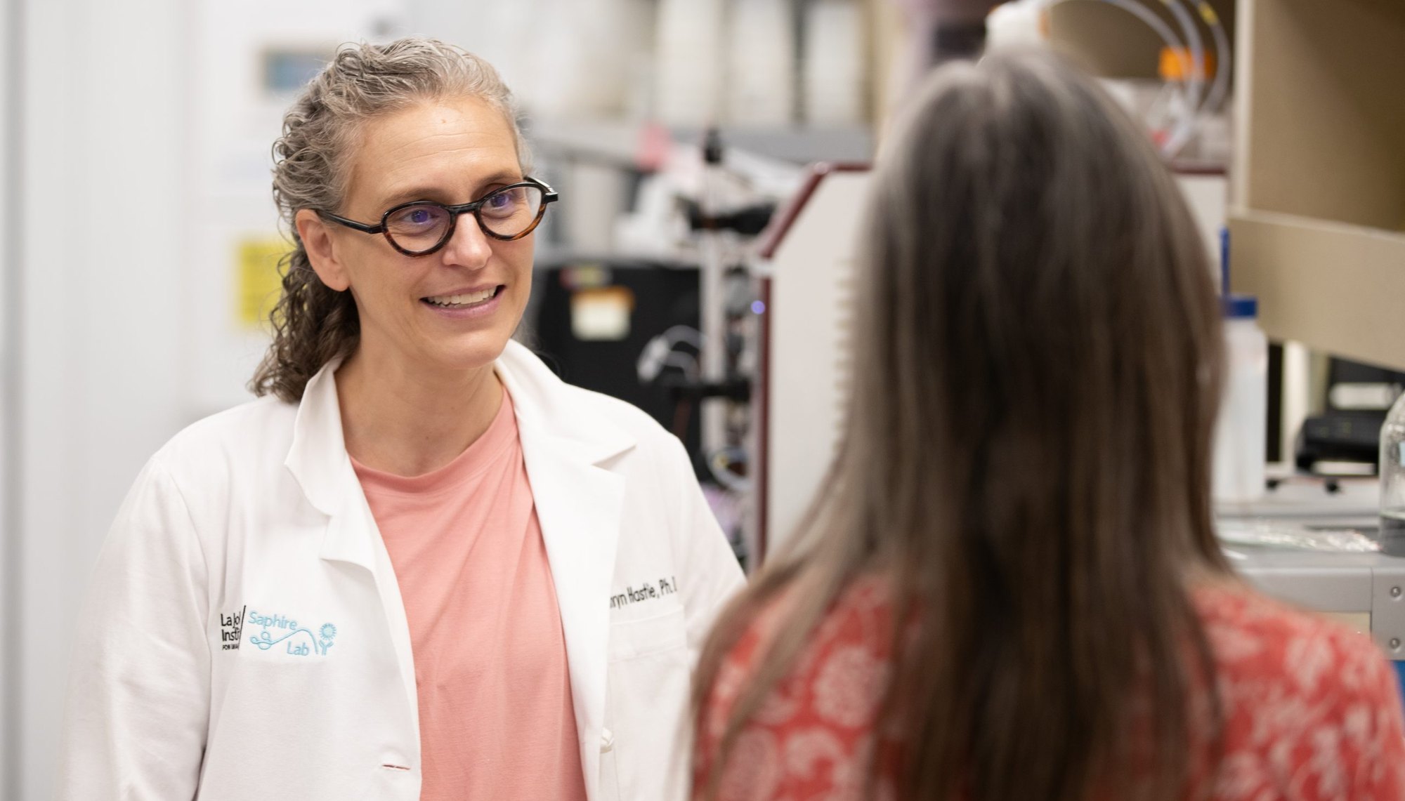 Researcher in white lab coat smiling inside a laboratory.
