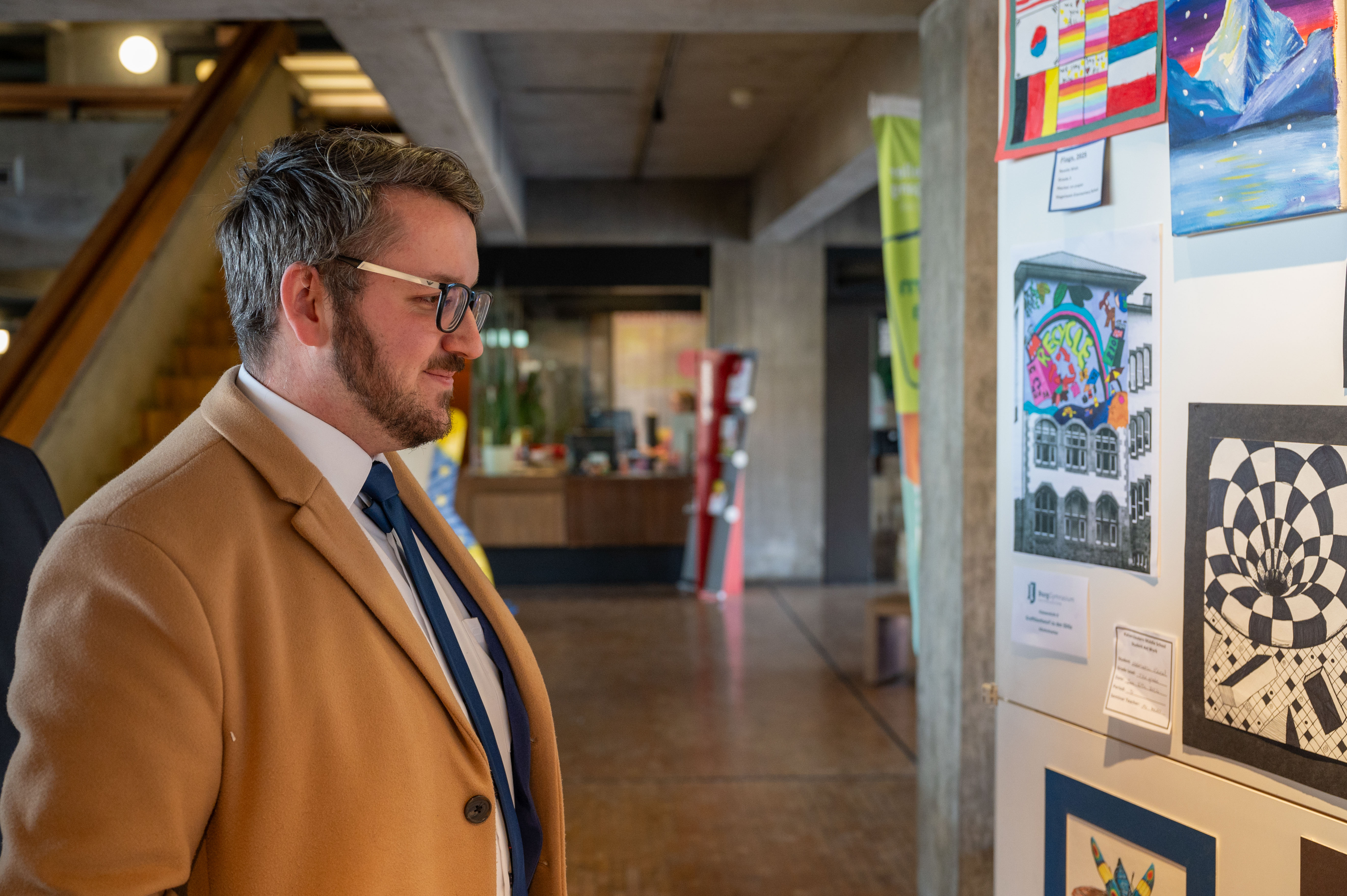 A man in a tan coat and glasses stands before a board covered in colorful student artworks.