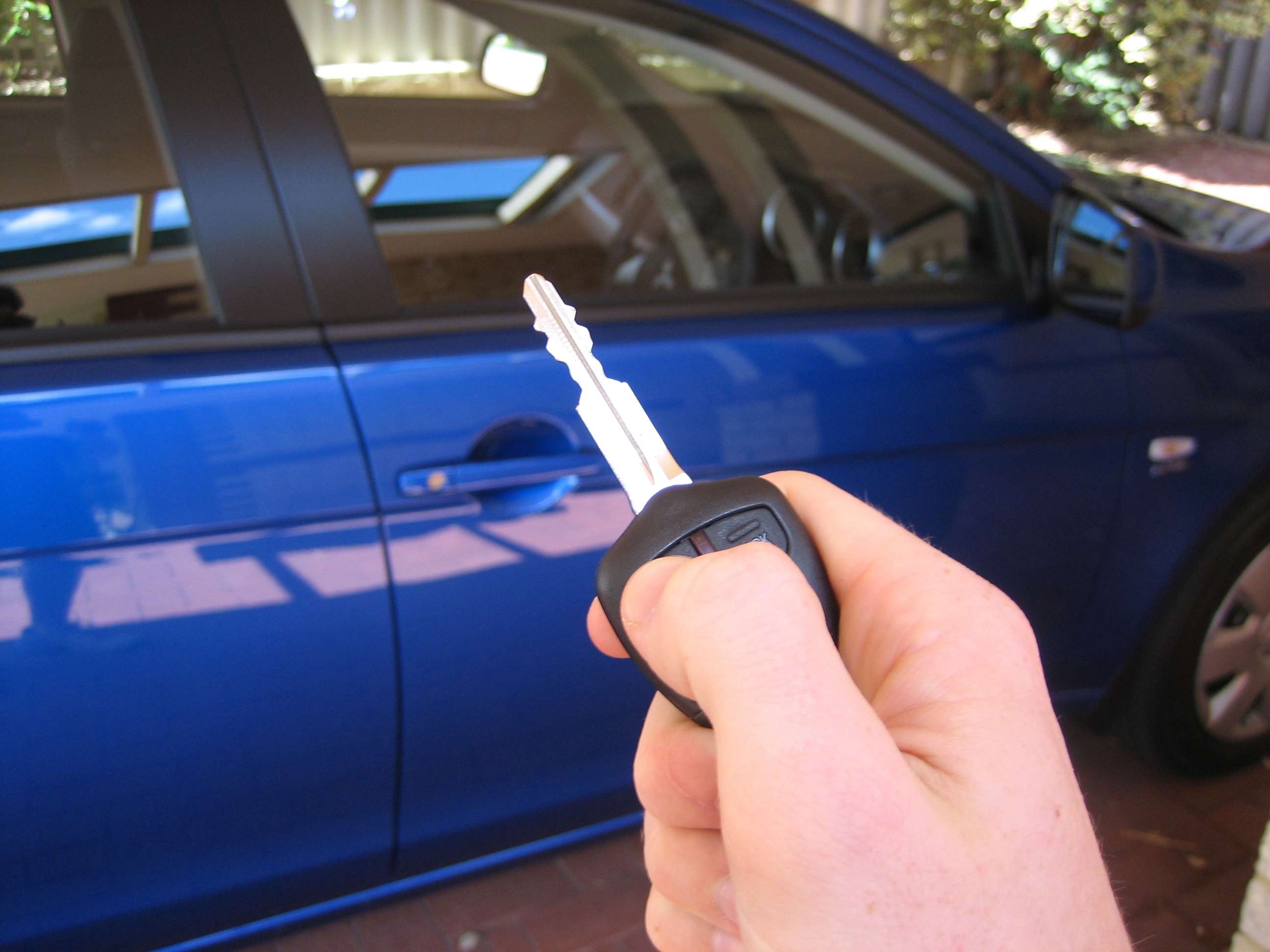 A hand holds a car key with a remote fob, a blue sedan visible behind it.