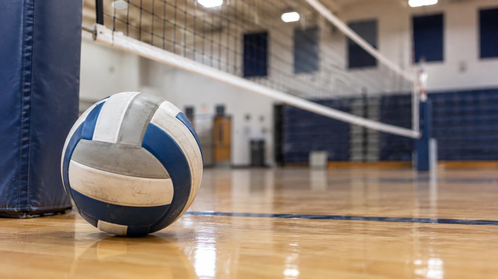 A volleyball resting on a hardwood gym floor near a volleyball net.