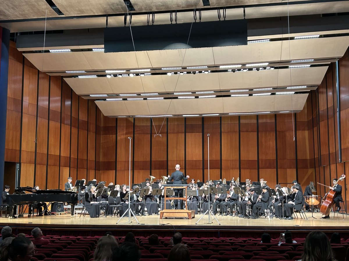 Large wind ensemble in black attire performing on a concert hall stage with conductor on podium.