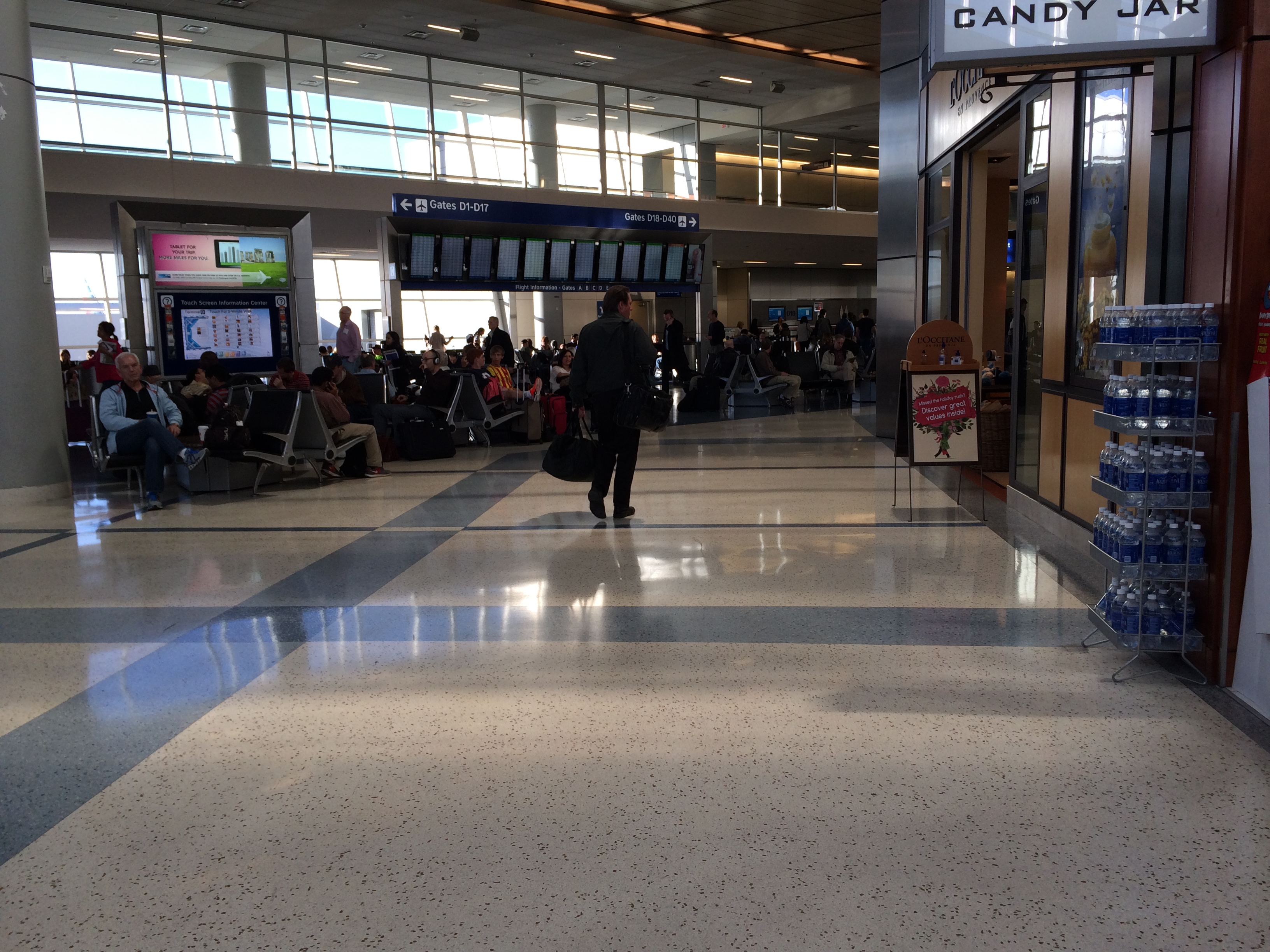 Airport terminal concourse with gate signage, travelers, and retail shops on a polished floor.
