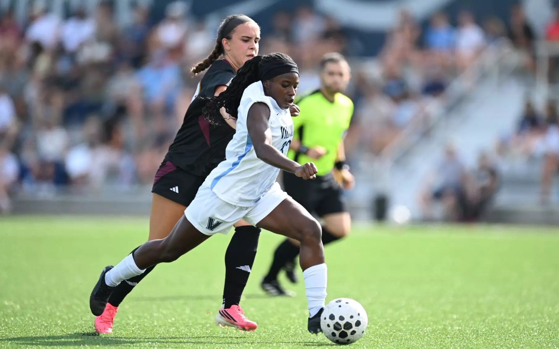 A Villanova player in white dribbles past a defender in a dark uniform on a grass field.