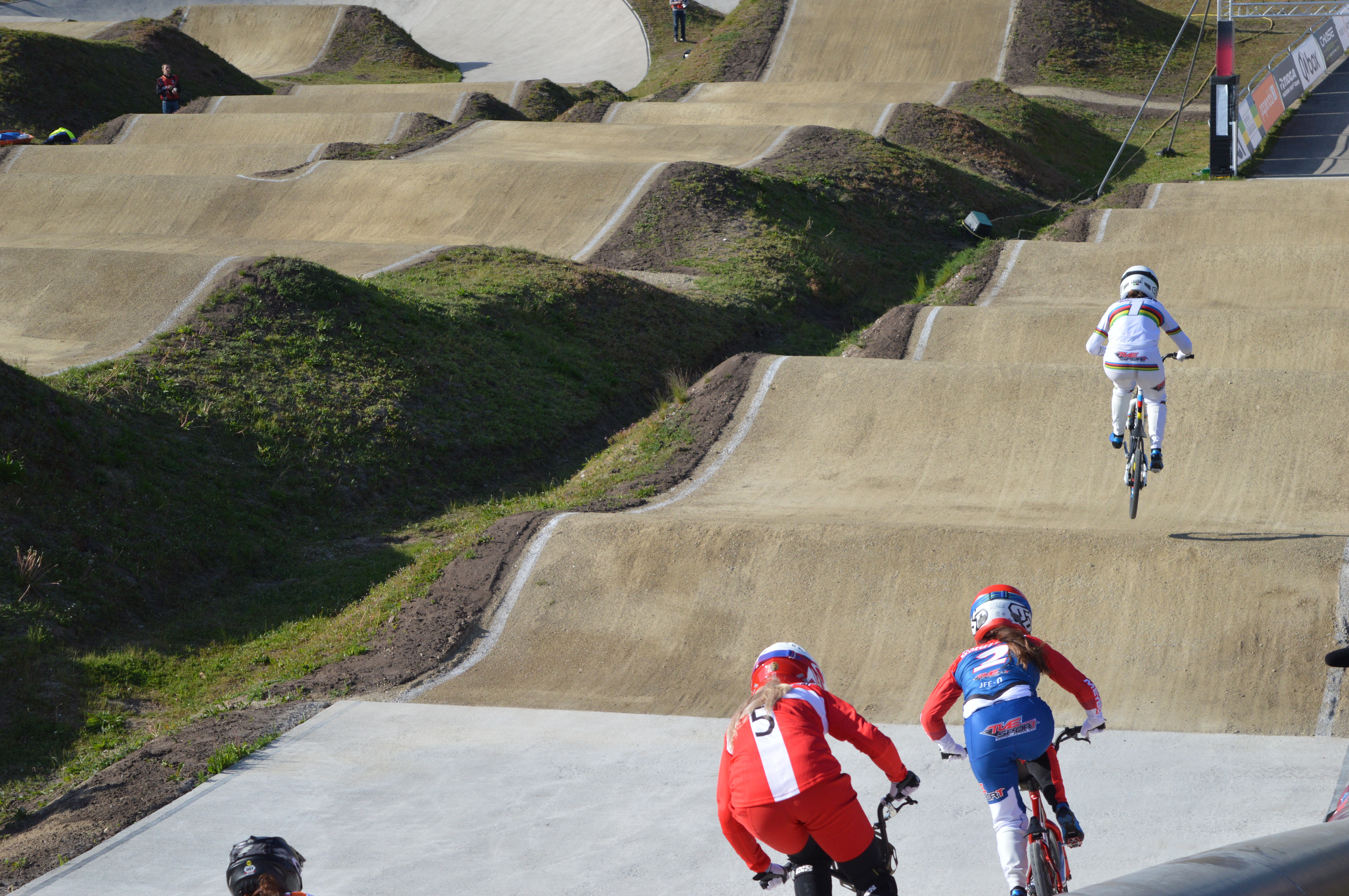 Four BMX riders race on a dirt track with rollers and berms.