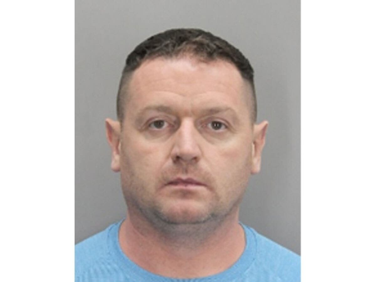 Mugshot of a middle-aged man with short dark hair and stubble wearing a light blue t-shirt.