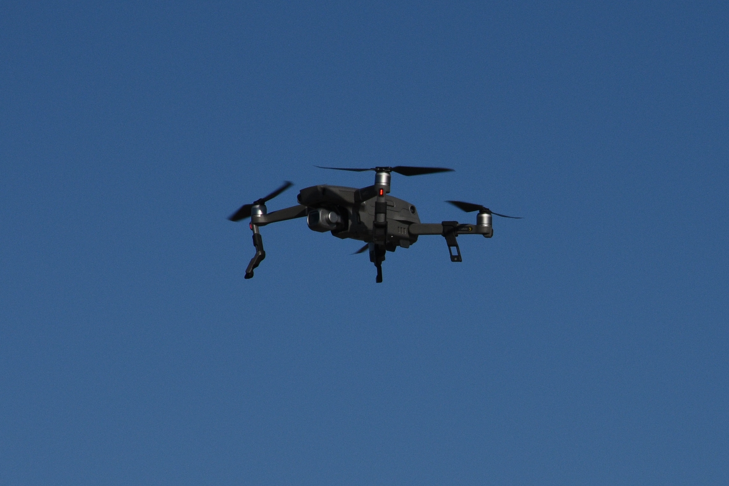A dark gray quadcopter drone with camera flying against a clear blue sky.