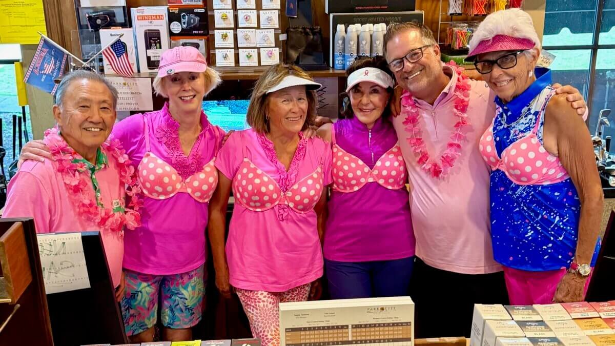 Group of seven people in pink clothing and accessories posing together indoors at a golf club.