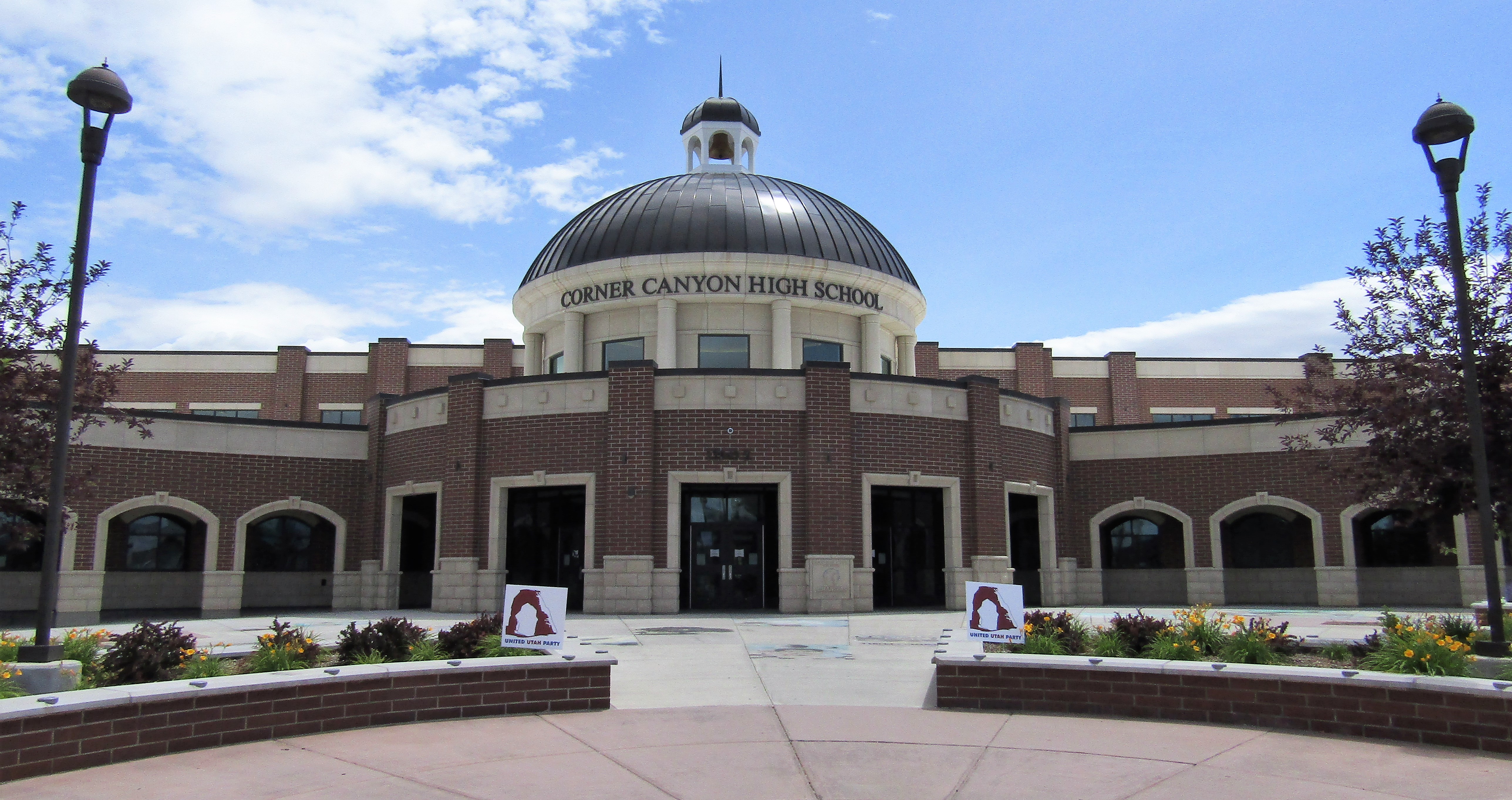 Brick school building with a domed entrance, lamp posts, and landscaped plaza under a partly cloudy sky.