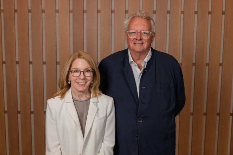 A woman in a white blazer and a man in a navy blazer posing together in front of a wood-paneled wall.