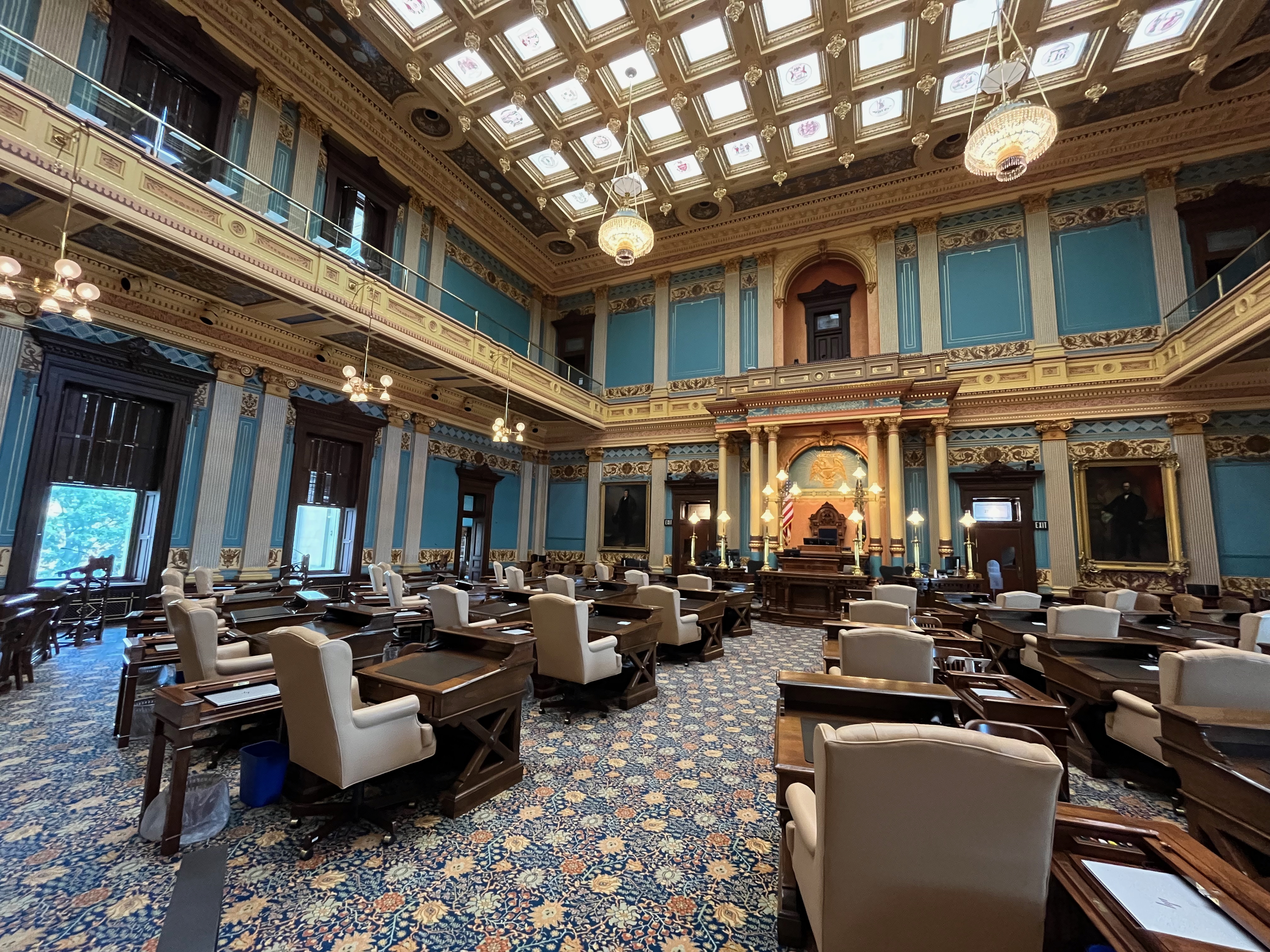 Empty legislative chamber with wooden desks, ornate gold walls, and a glass skylight ceiling.