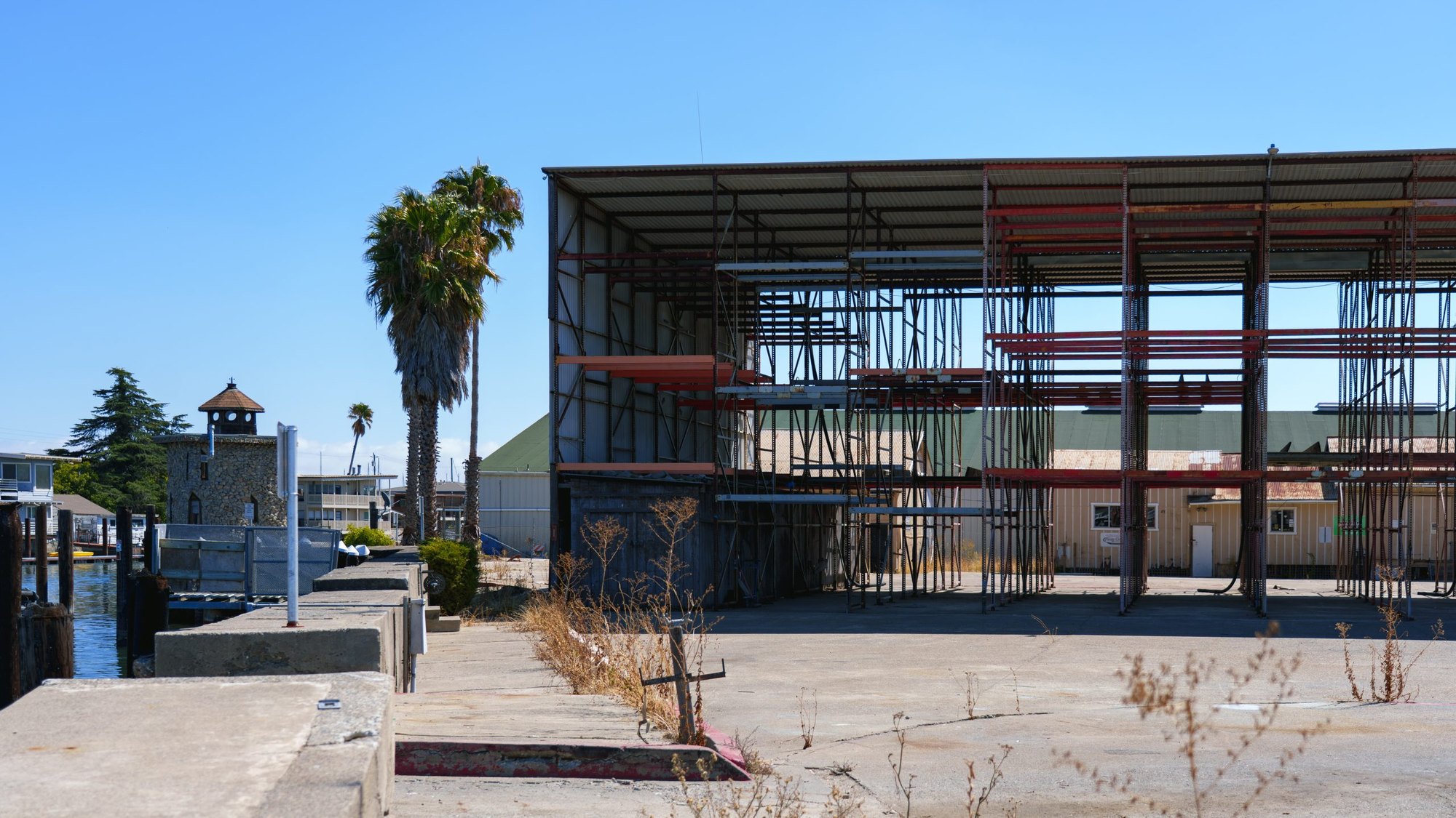 Steel boat storage rack on a concrete waterfront lot with a dock and palm tree in the background.