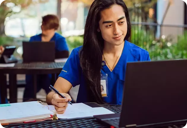 A Chamberlain University nursing student in blue scrubs writes in a binder while studying on a laptop at a table.