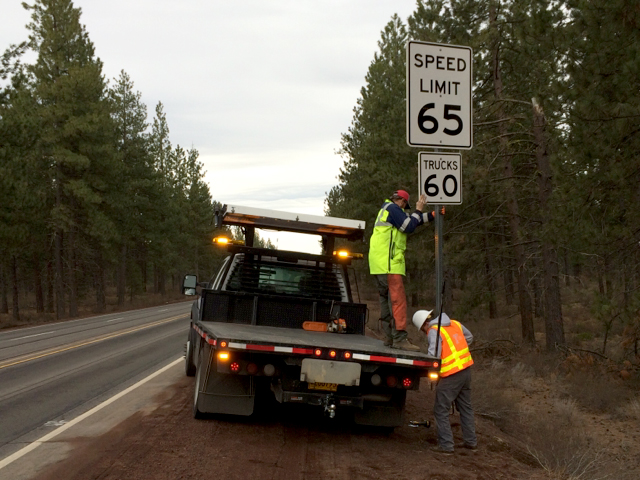 Two workers in high-visibility vests installing a speed limit sign on a roadside post.