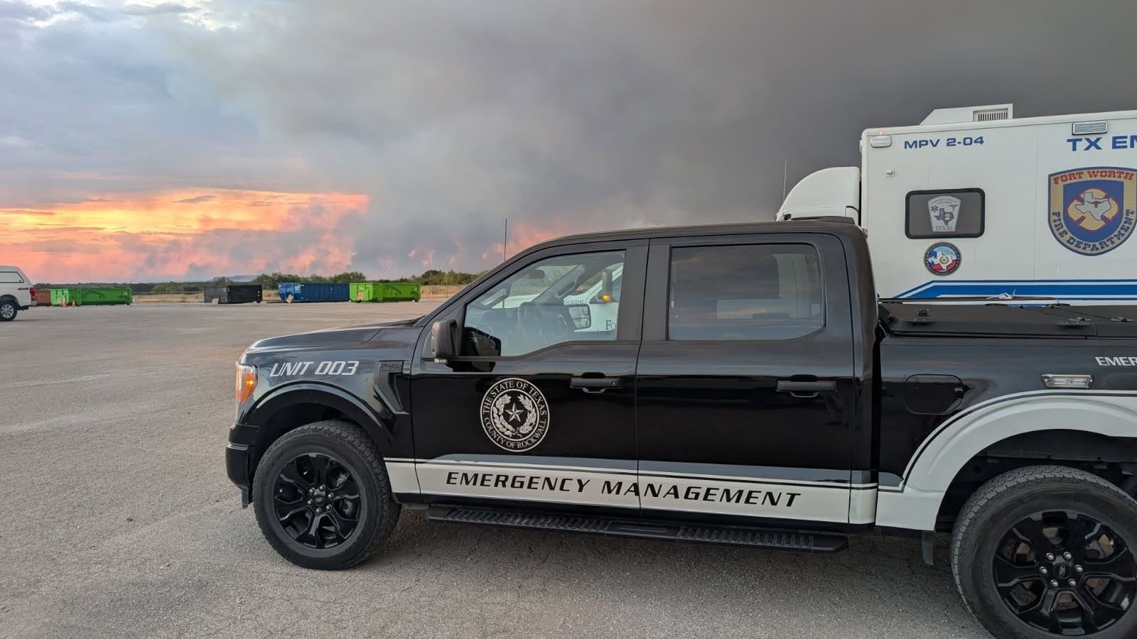 Black pickup truck marked Emergency Management parked near a large wildfire smoke plume.