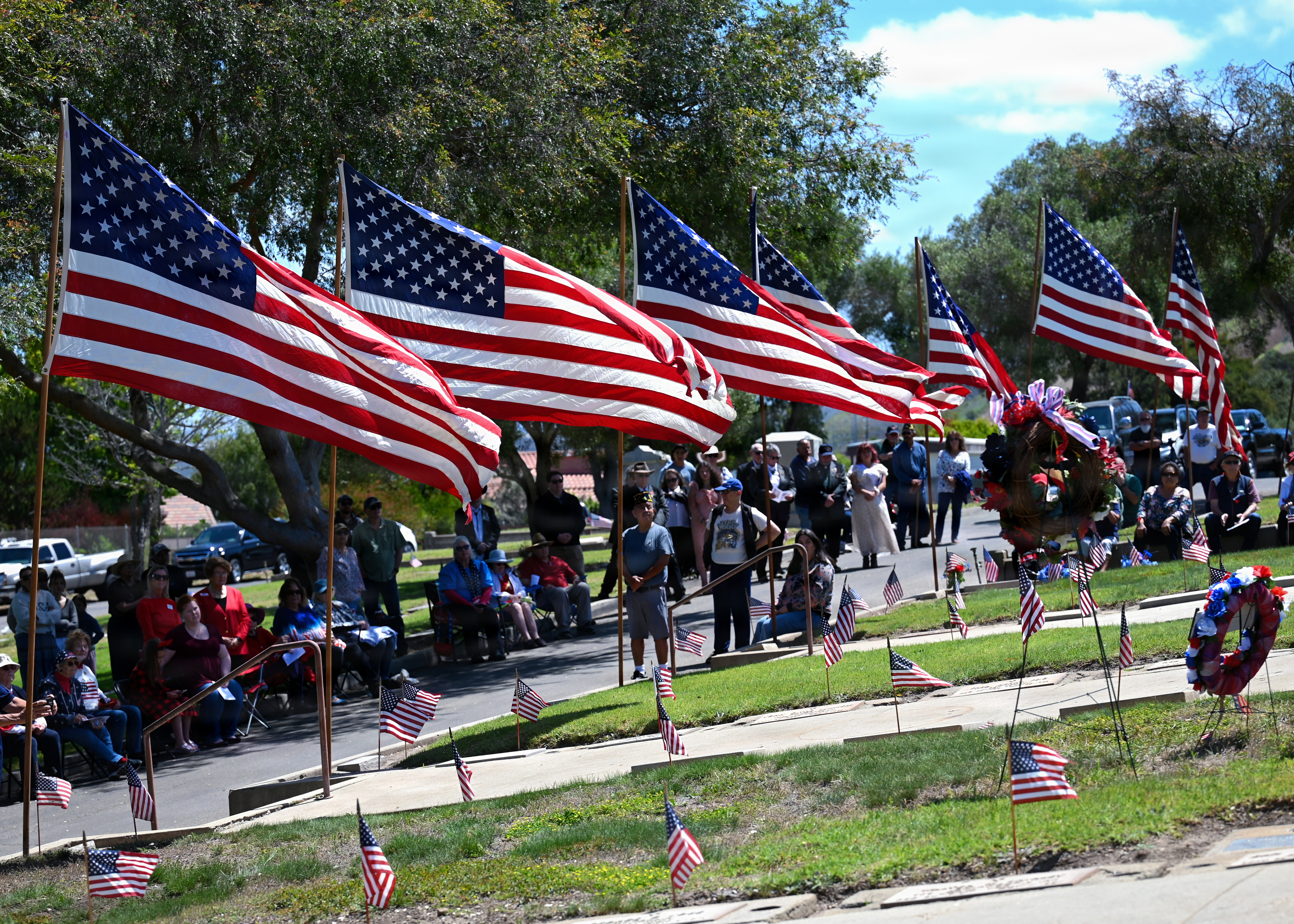 Large American flags lining a cemetery walkway with a crowd gathered on both sides.