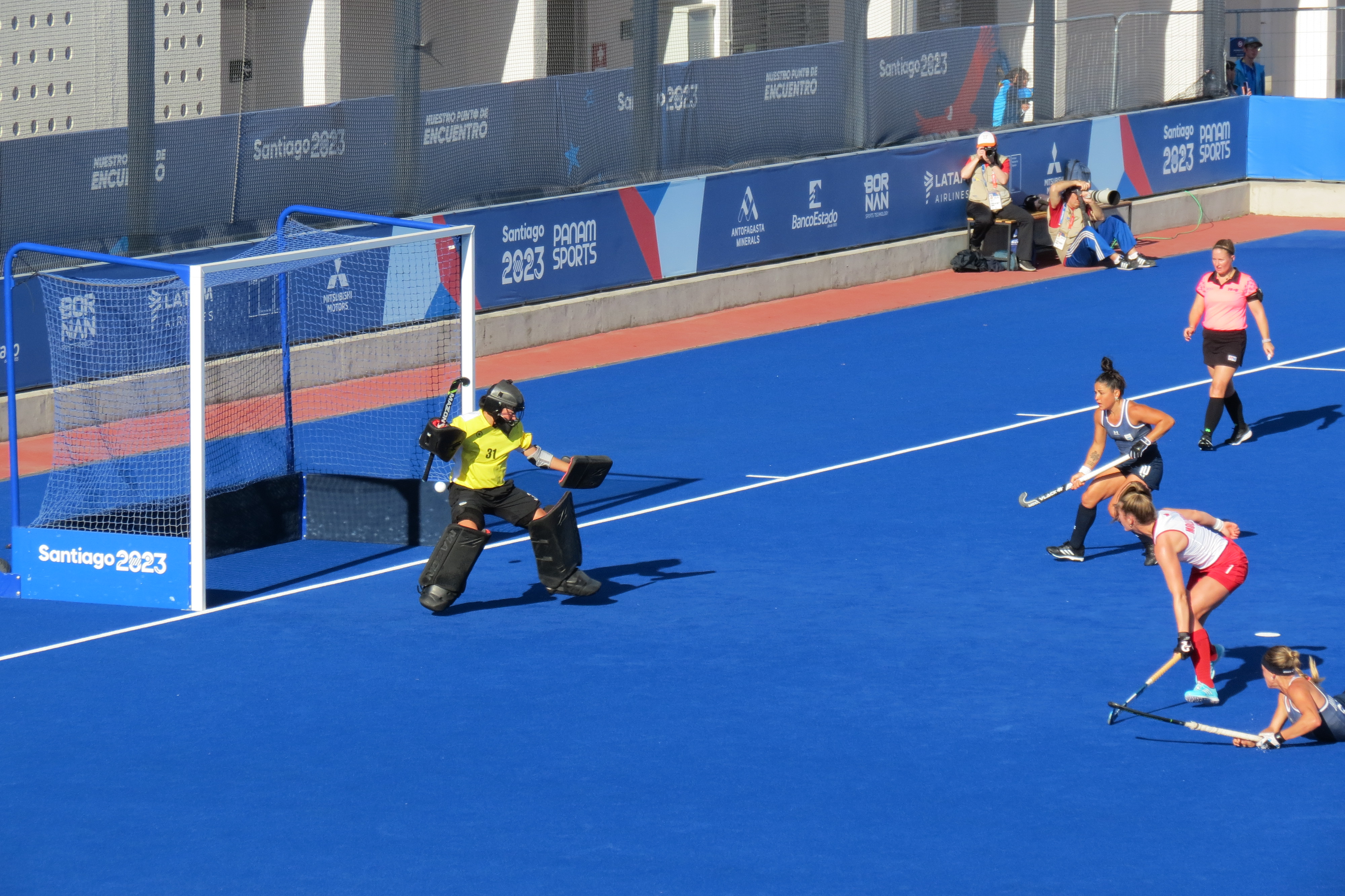 Women's field hockey players in action near a goal on blue turf.