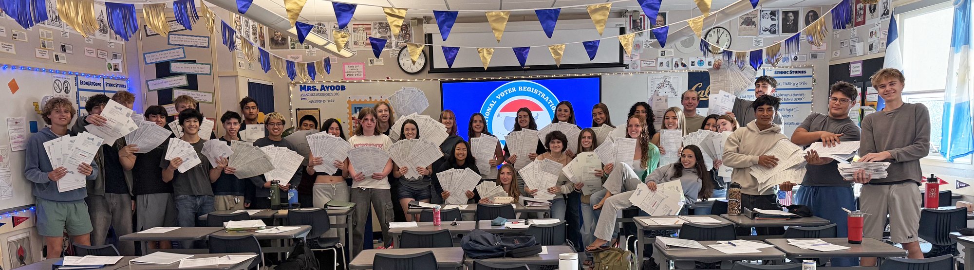 Students standing in a decorated classroom each holding up papers.