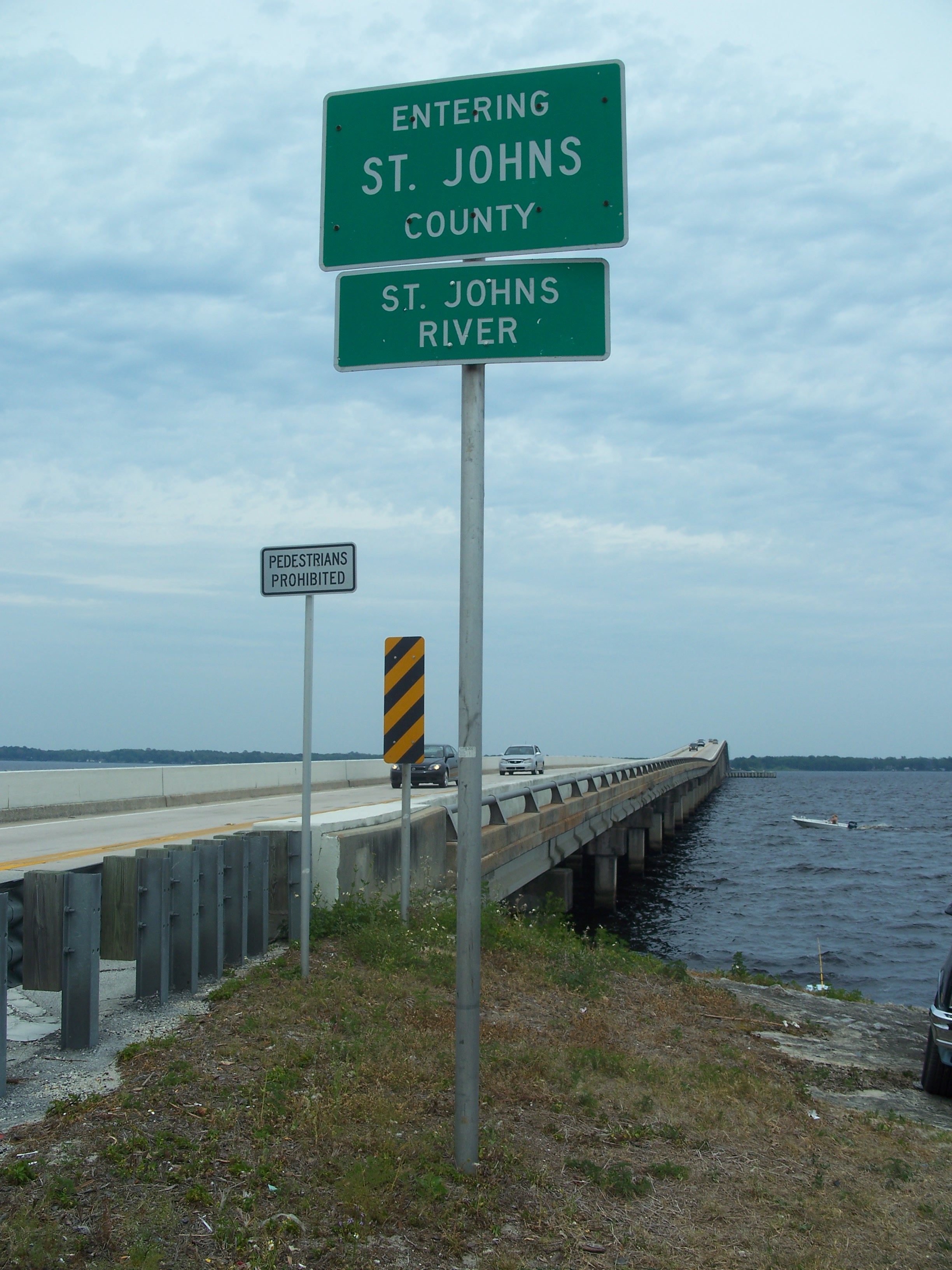Highway signs mark St. Johns County and the St. Johns River beside a long bridge over open water.