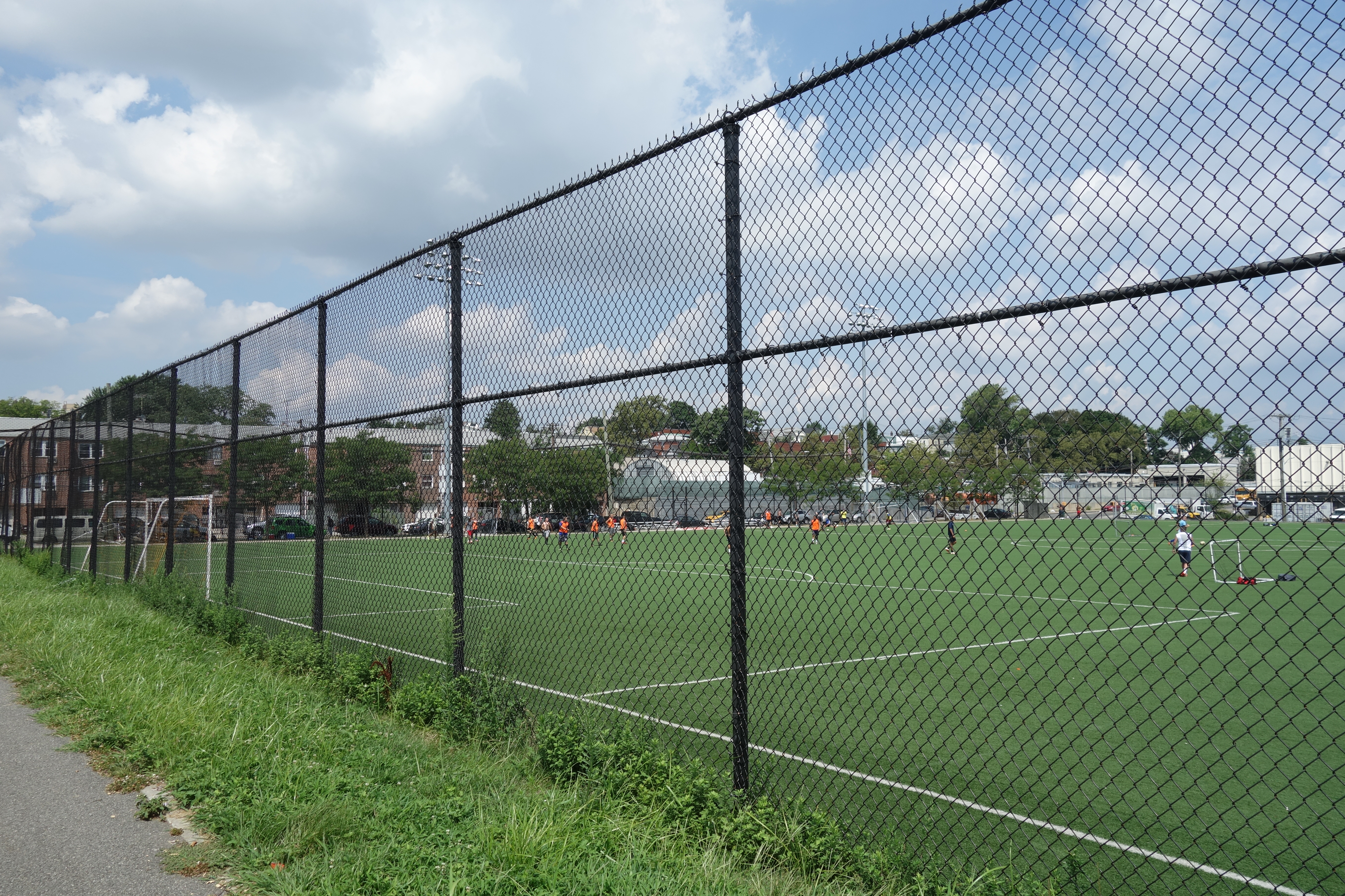 Players on a green artificial turf soccer field viewed through a chain-link fence.