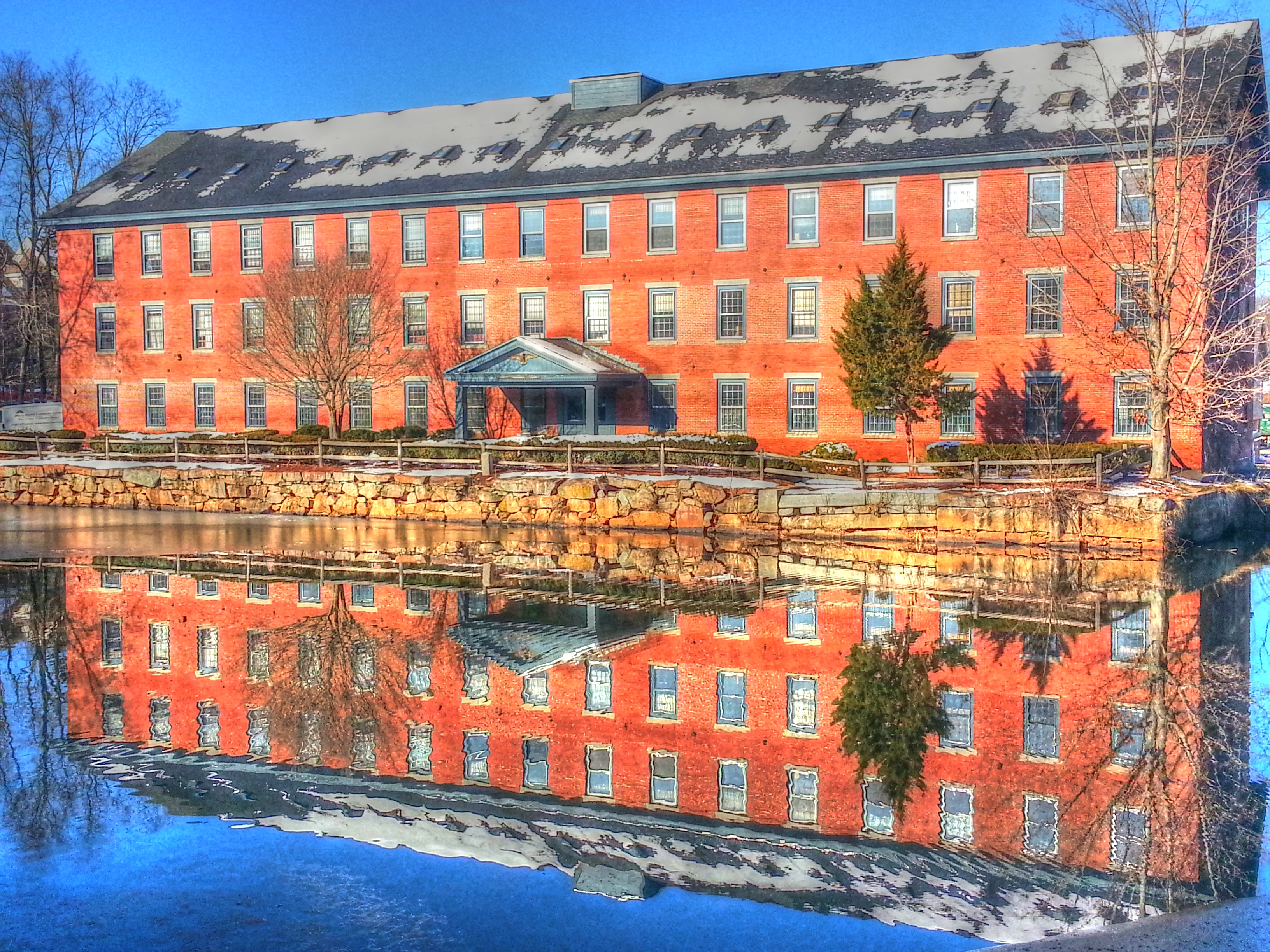 Four-story red brick mill building reflected in a still millpond, bare winter trees behind.