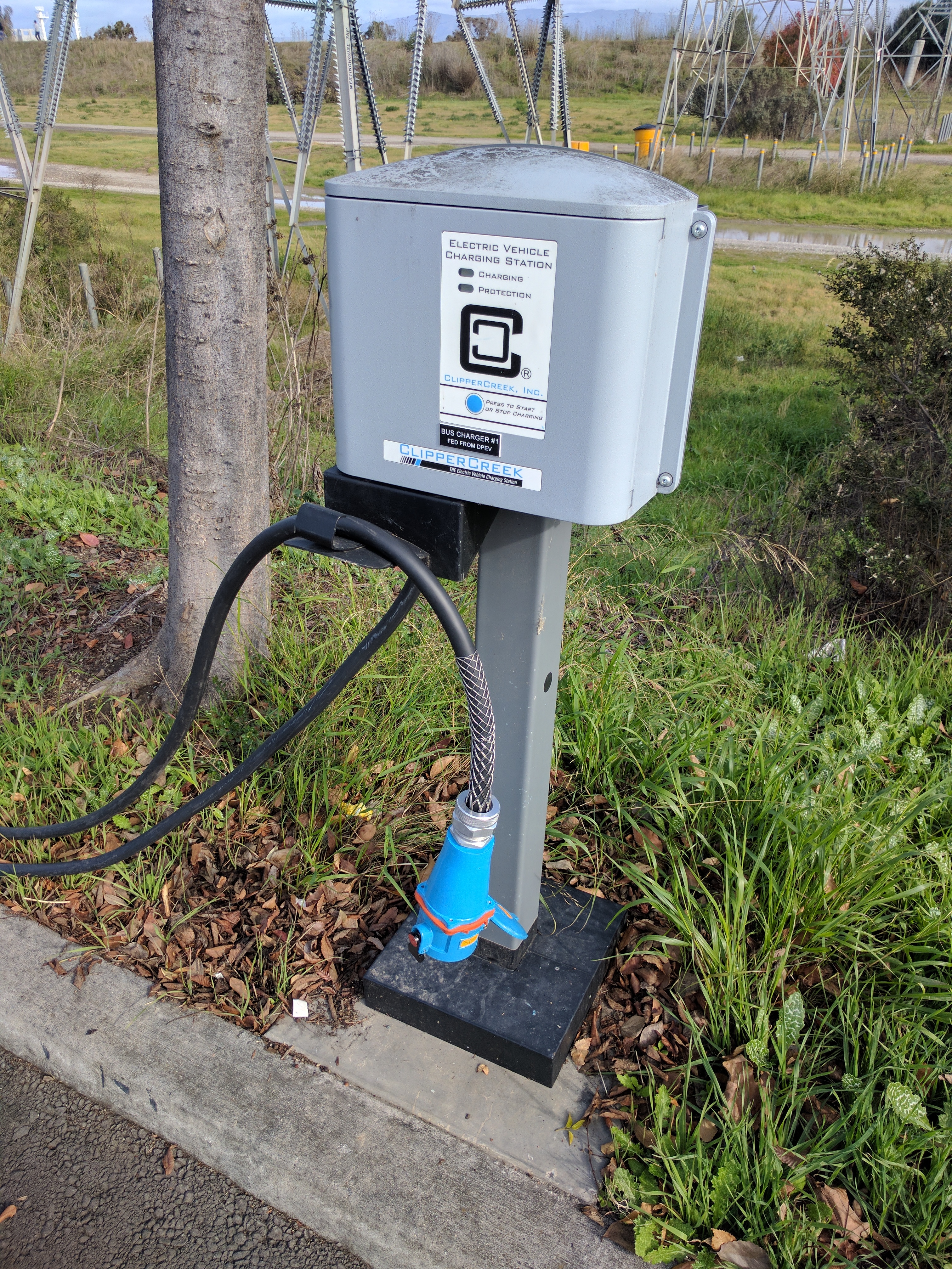 A gray metal electric vehicle charging station on a post with a black cable and blue connector plug.