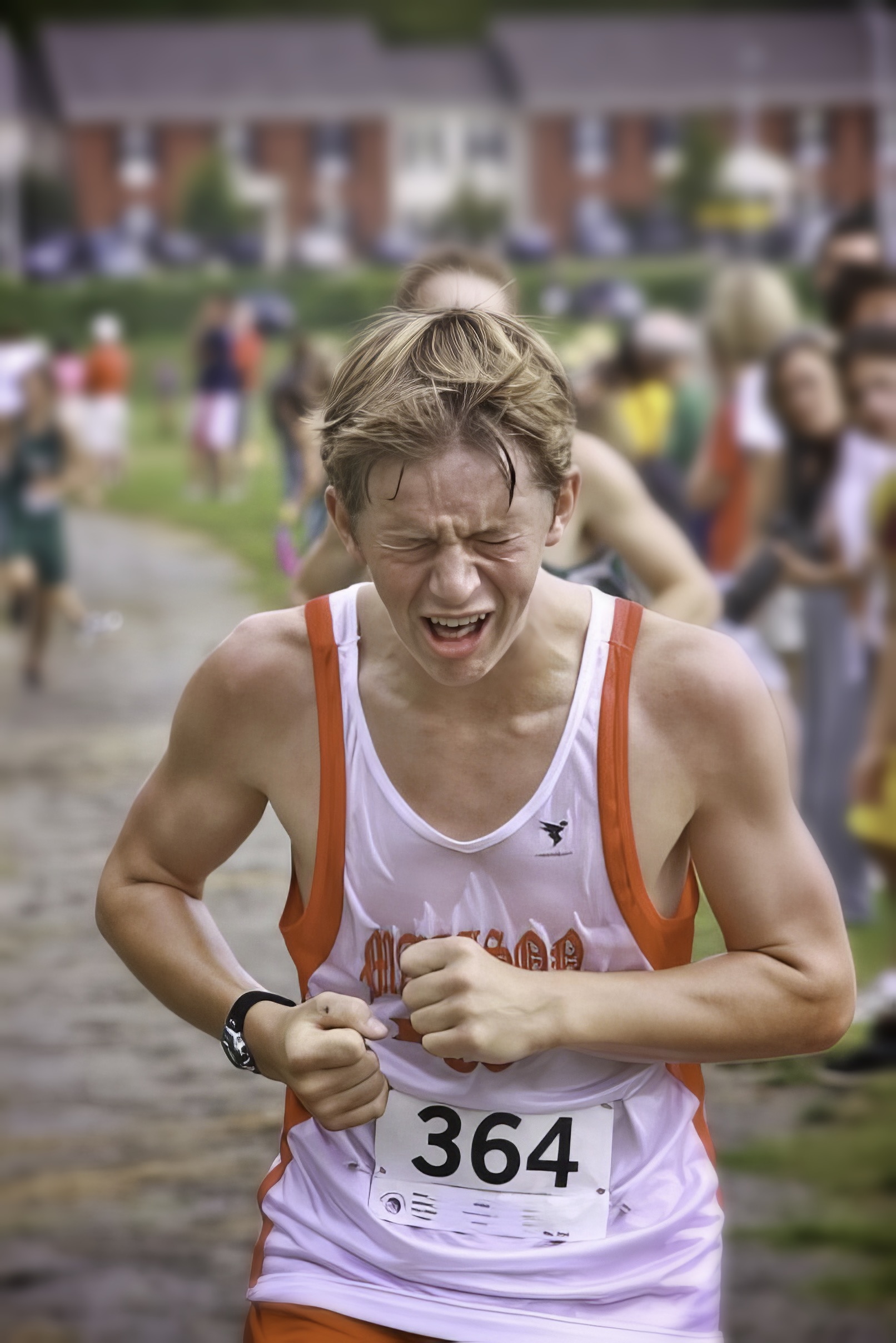 A male runner in a white and orange singlet grimaces with effort mid-race.